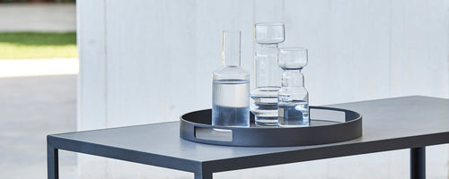 A round serving tray holding various glassware, including a tall pitcher and several glasses, placed on a sleek table.
