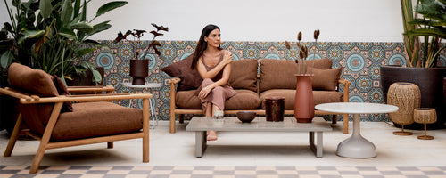 A stylish seating area featuring a brown sofa, a lounge chair, and a decorative table, surrounded by lush greenery and patterned decor.