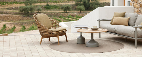 Modern seating area featuring a woven chair, a gray sofa, and two stylish coffee tables on a textured surface. Natural scenery in the background.