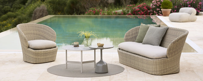 Stylish seating arrangement with two woven chairs, a round table, and decorative accents, set beside a serene pool.