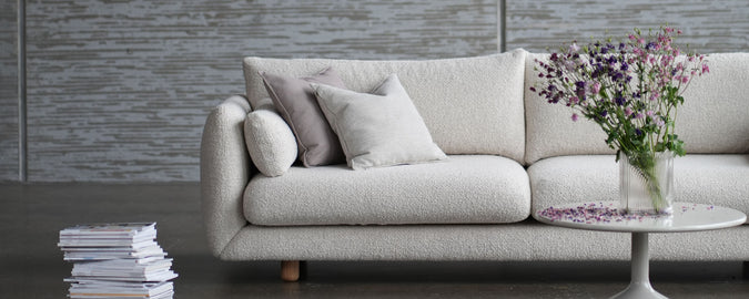 Stylish light-colored sofa with decorative pillows, accompanied by a small table featuring a vase of flowers and a stack of magazines.