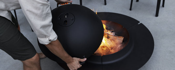 A person is lifting a large black dome from a fire pit, revealing roaring flames and burning logs inside.