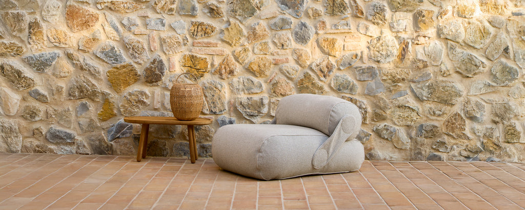 Beige lounge chair next to a wooden table against a stone wall.