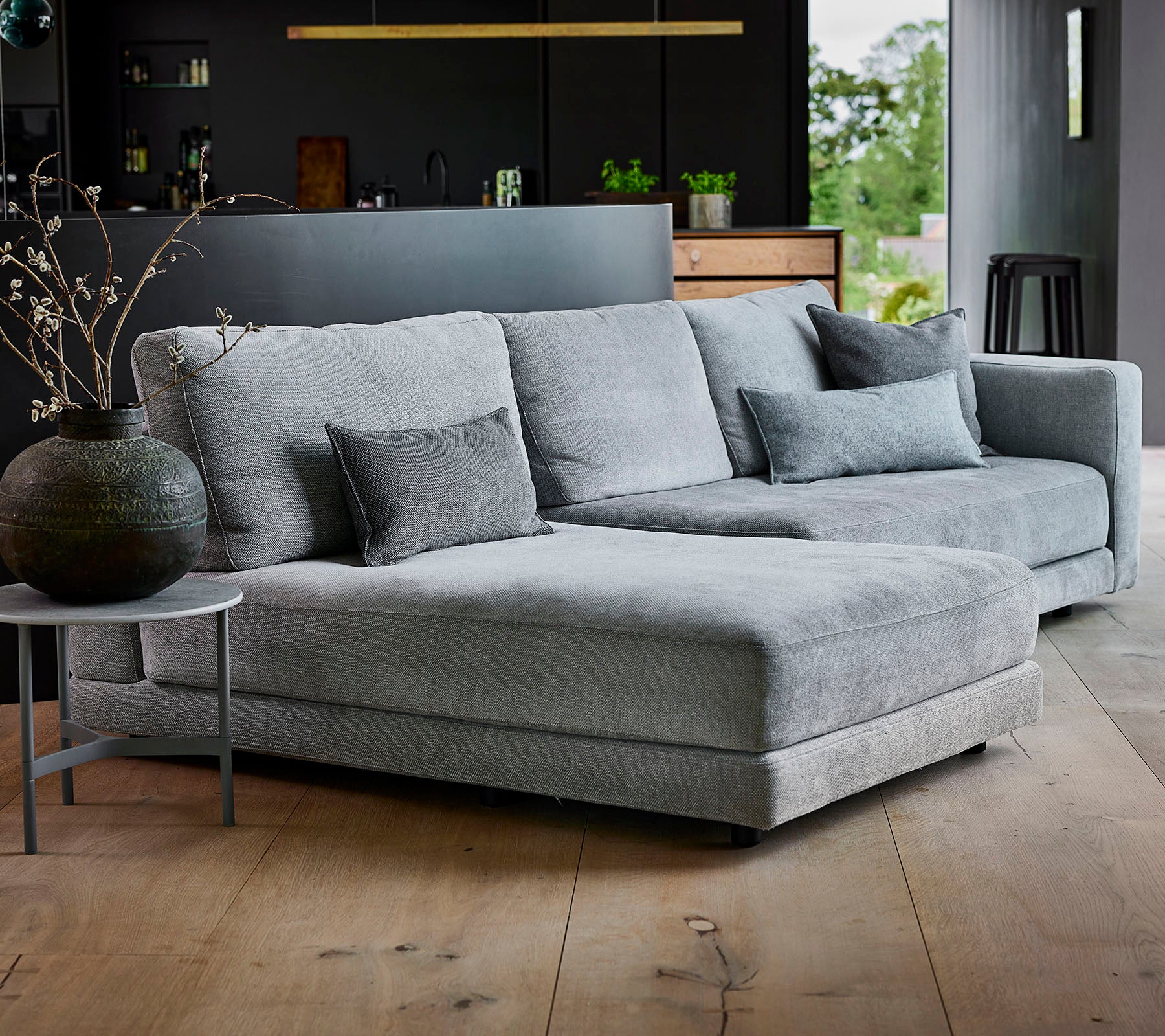 Grey sectional sofa with textured fabric and decorative cushions, accompanied by a round side table and a stylish vase.