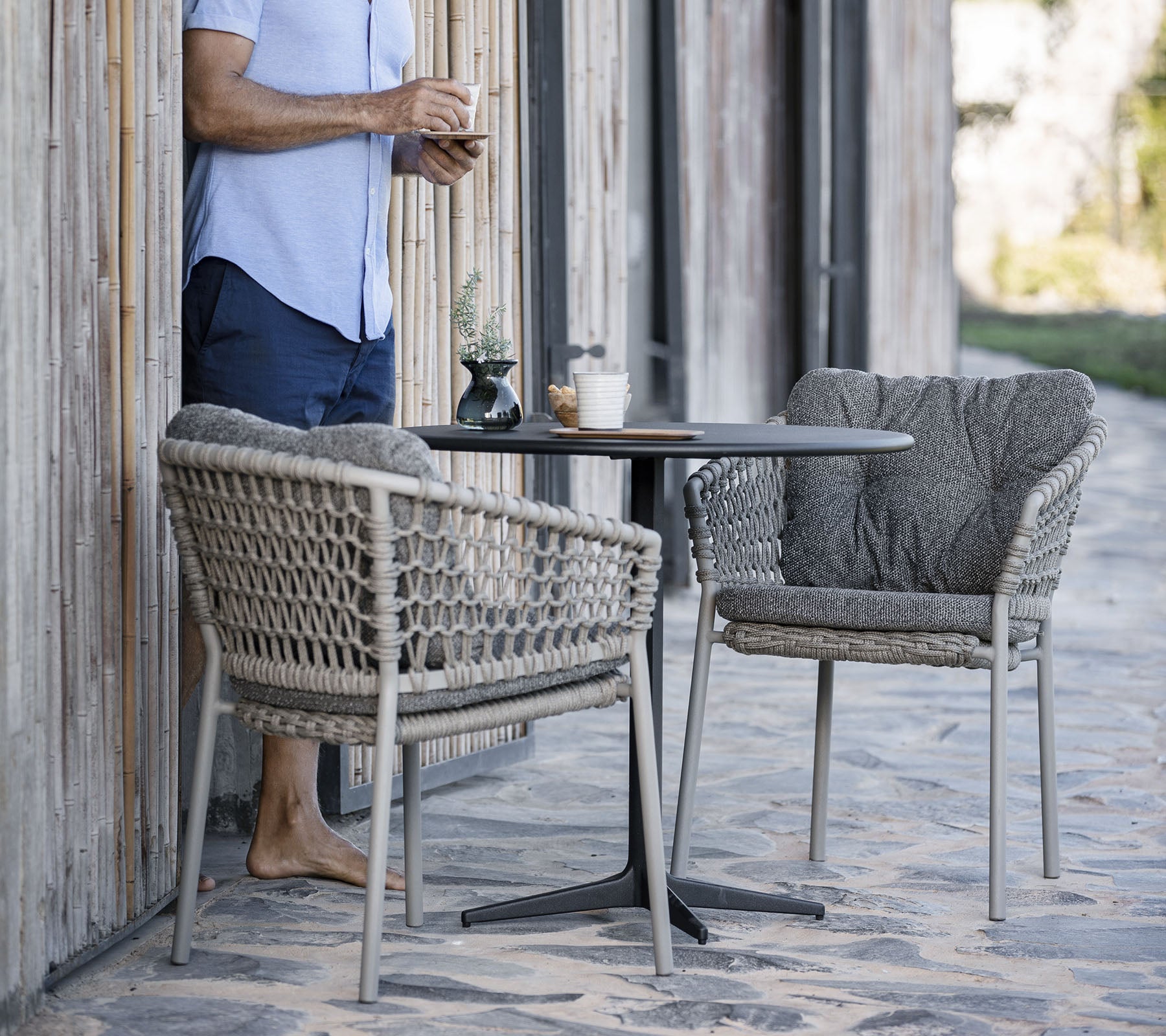 A person stands beside a small round table with two stylish woven chairs, enjoying a drink in a relaxed setting.