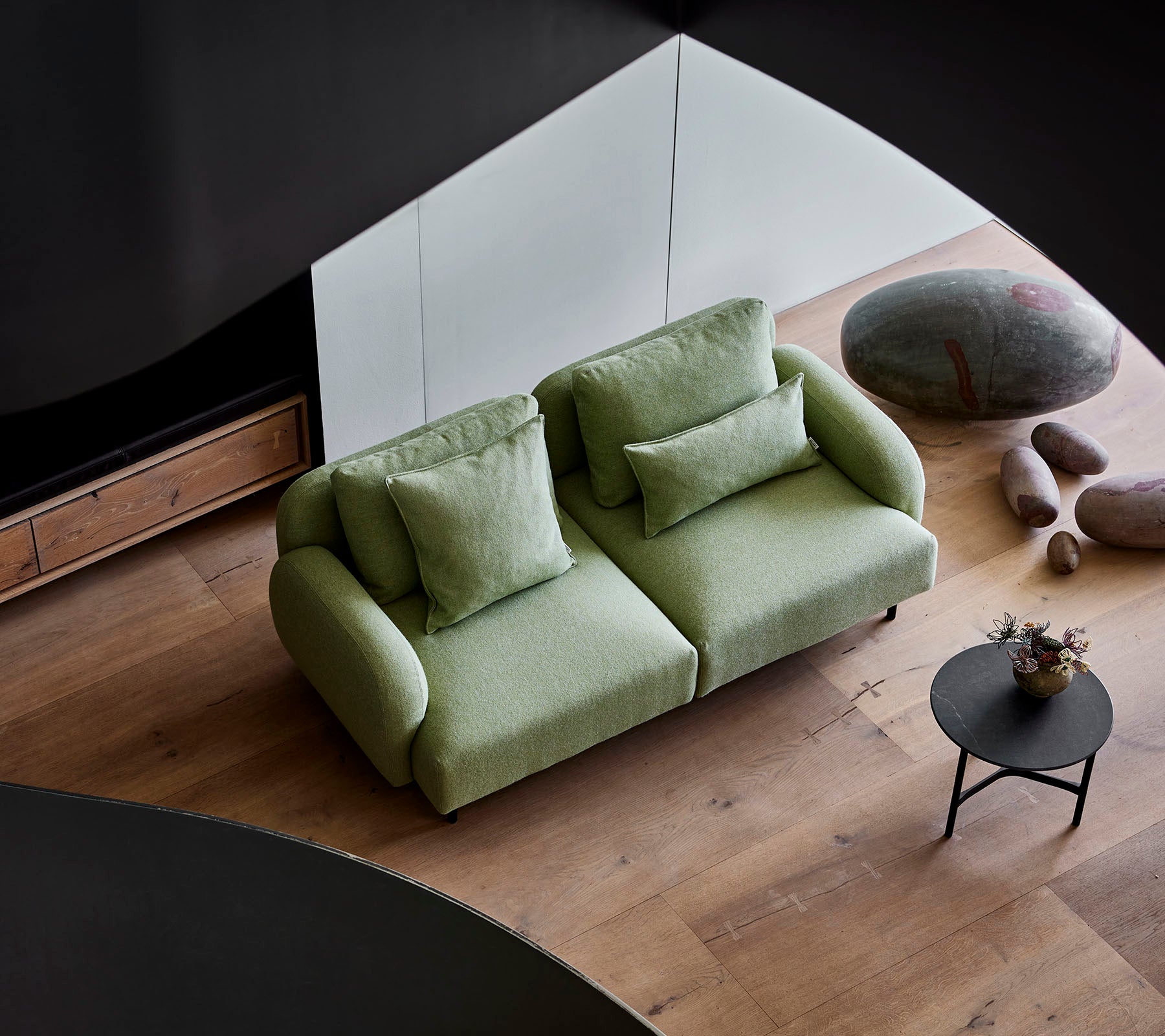 Stylish green sofa with plush cushions beside a small round table, set against a modern backdrop featuring wooden flooring and smooth stones.