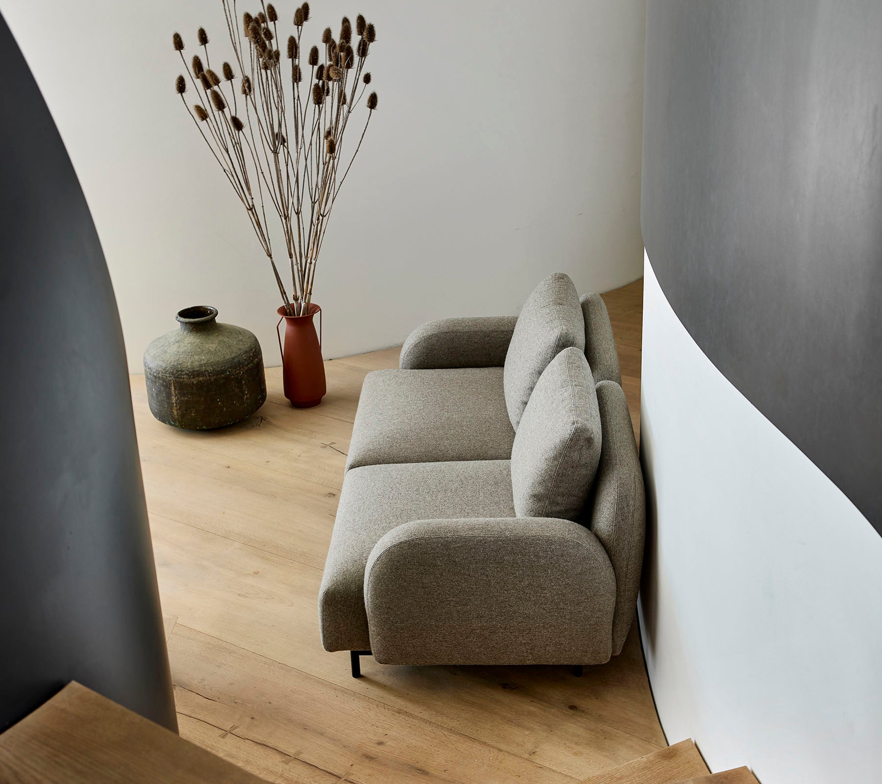A cozy beige sofa positioned beside a decorative vase and dried plants, set against a curved wall and wooden flooring.