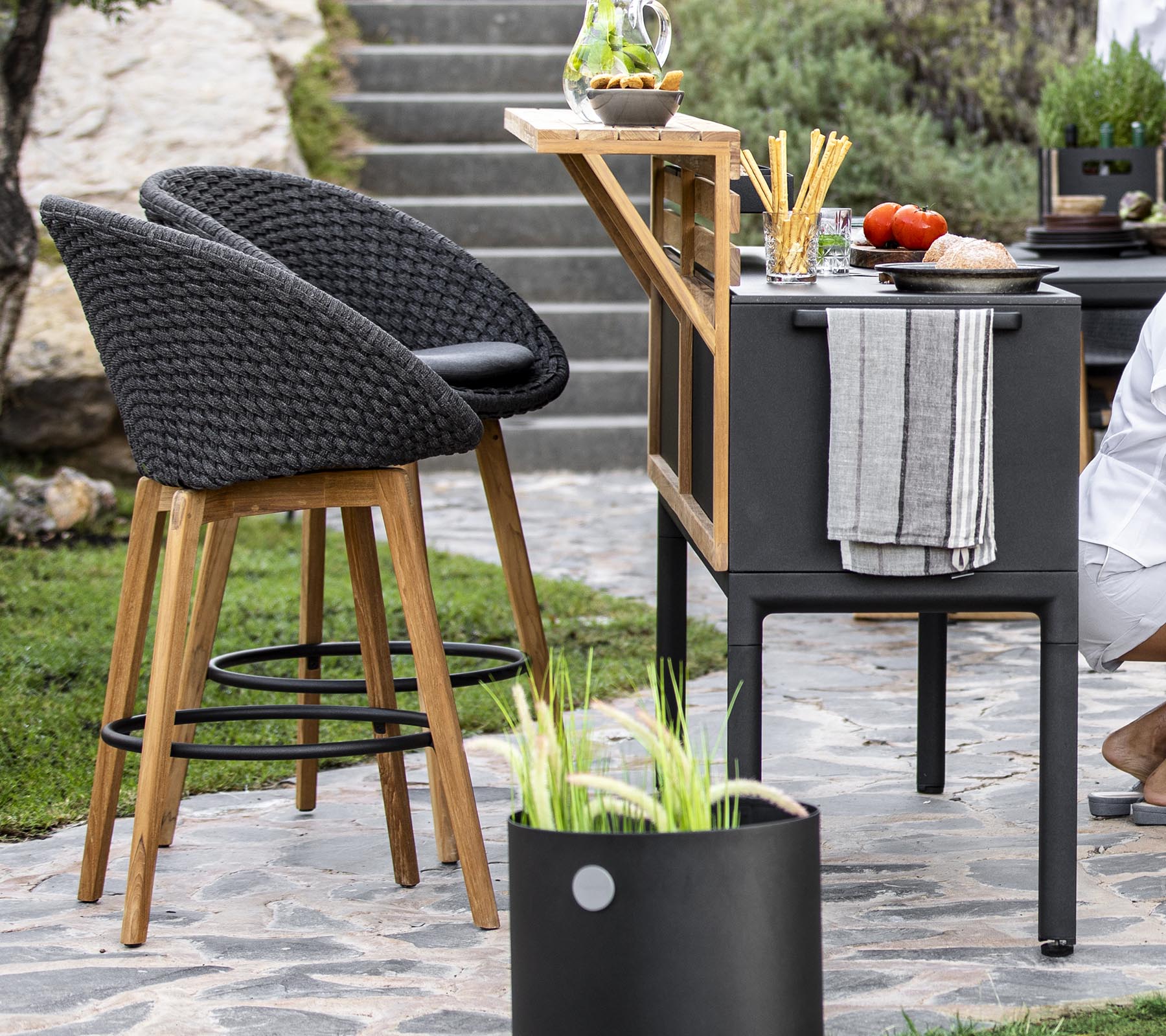 Stylish outdoor seating with wicker chairs, a modern bar cart, and fresh ingredients arranged on a countertop, complemented by decorative plants.