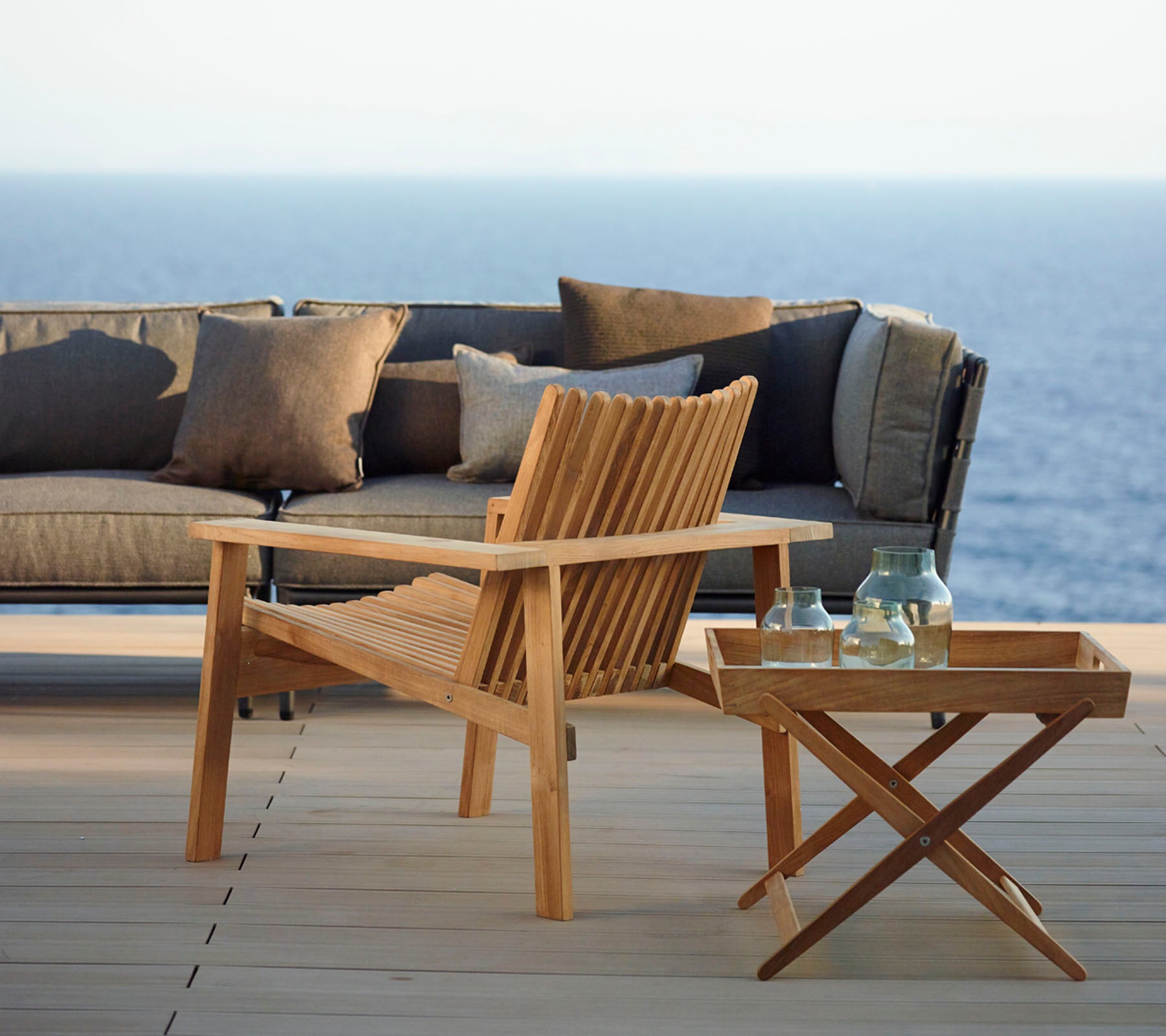 Stylish wooden chair and side table with bottles, accompanied by a modern sofa adorned with cushions, set against a tranquil sea backdrop.