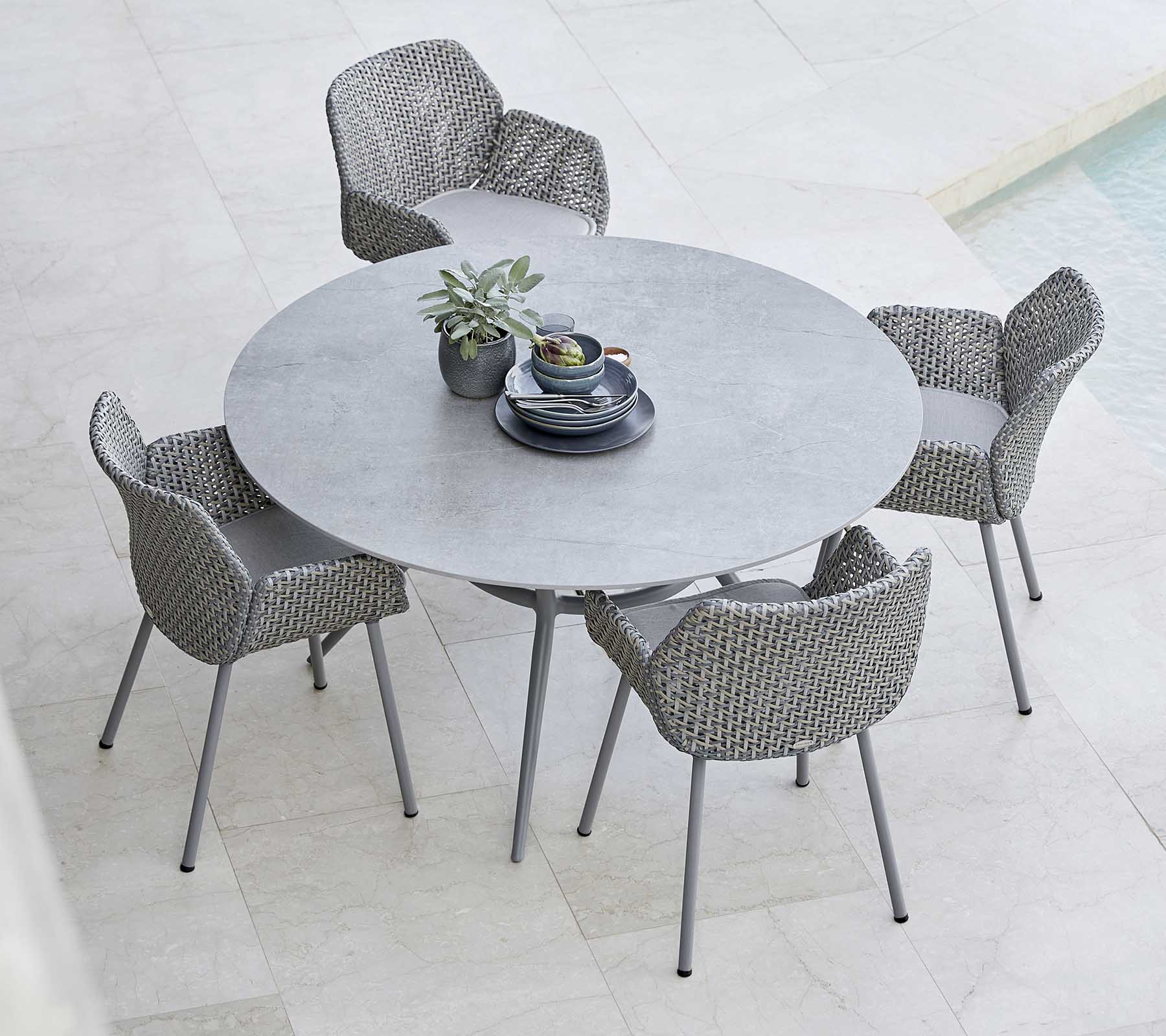 A stylish round table paired with woven chairs, featuring a plant and tableware in the center, perfect for outdoor dining.