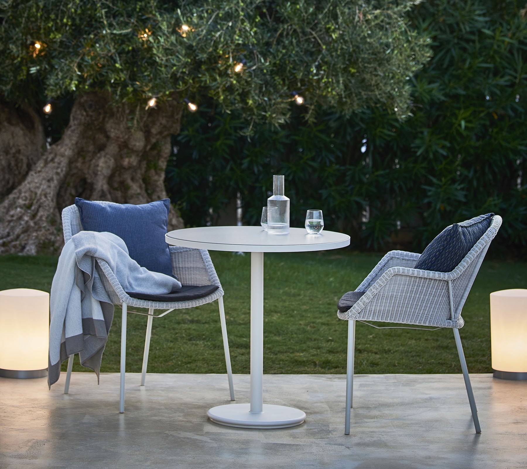 A small round table with two stylish chairs, a bottle of water, and a glass, set amidst lush greenery and softly glowing lights.