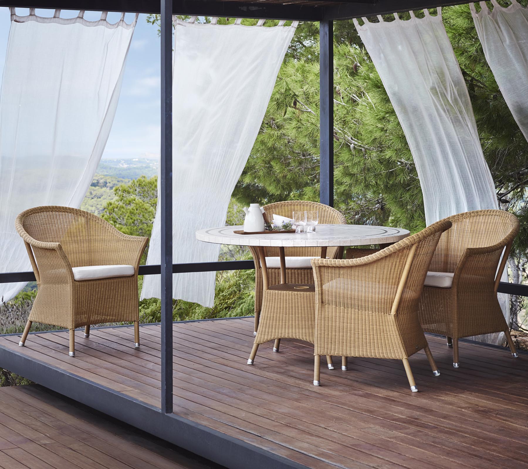 A stylish seating area features four wicker chairs and a round table, surrounded by sheer curtains and natural greenery.