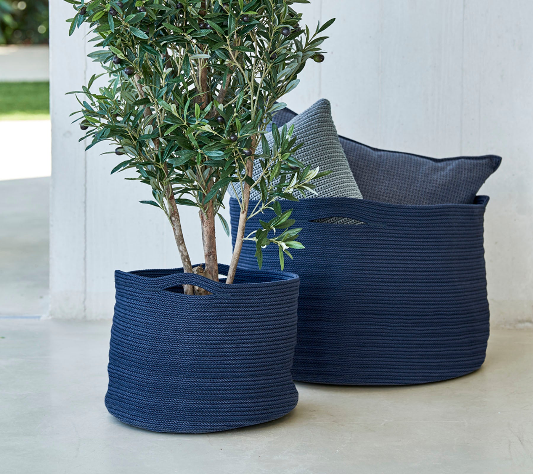 Two navy blue woven planters, one featuring a small tree, accompanied by a decorative pillow.