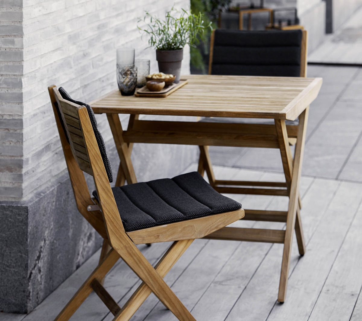 Wooden table and chair set featuring black cushions, adorned with potted plants and decorative glassware. Perfect for a cozy setting.