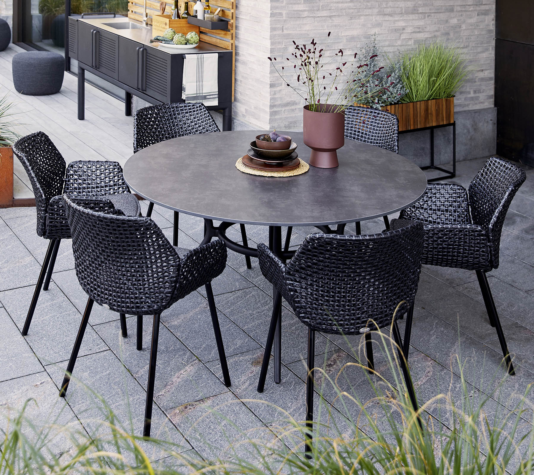 Round table with six black woven chairs, featuring a decorative bowl and a potted plant, set among greenery and modern decor.