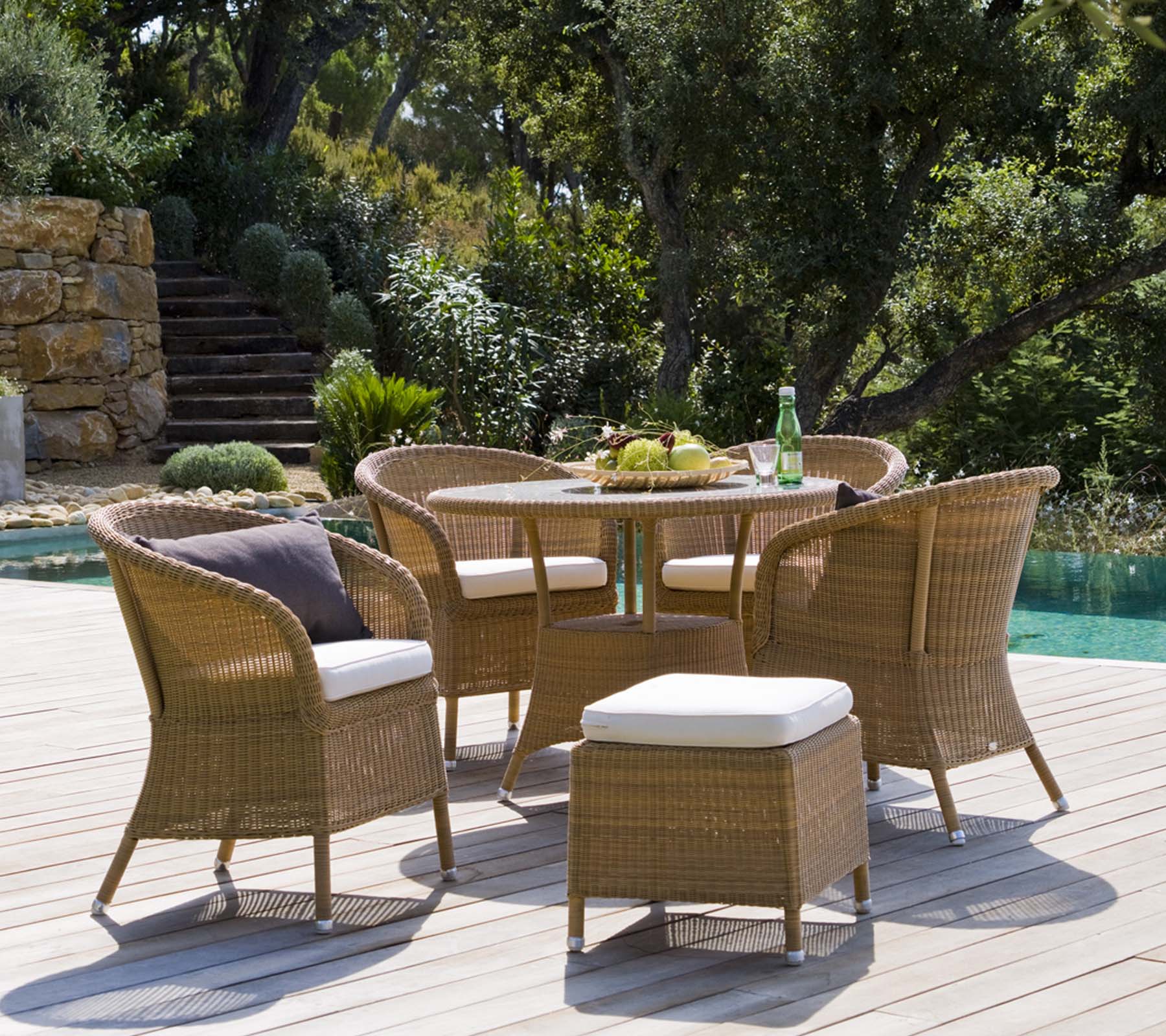 A stylish outdoor seating set featuring a round table, four woven chairs, and a pouf, surrounded by lush greenery and a tranquil setting.