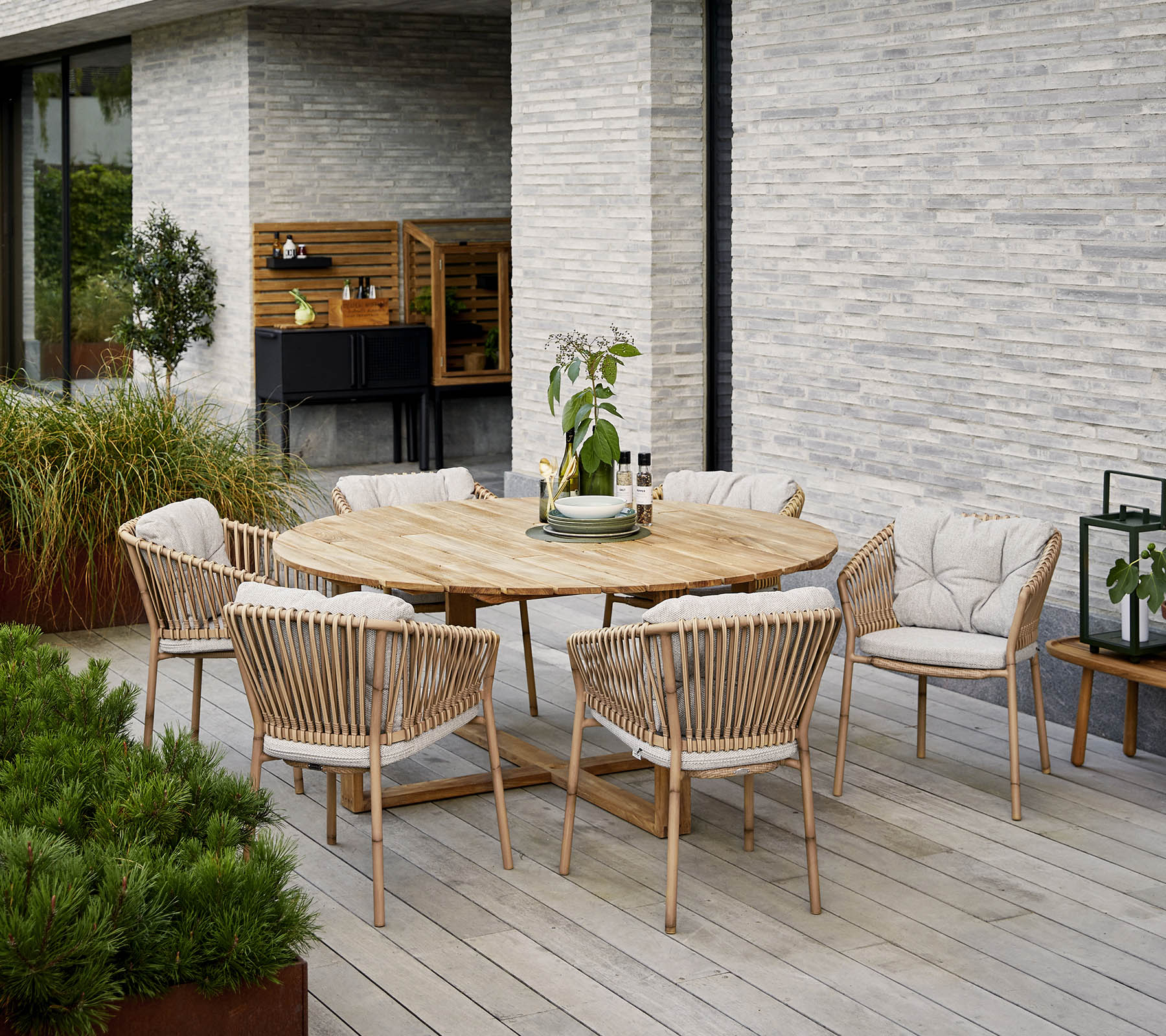 Round wooden dining table surrounded by upholstered chairs, with a decorative plant centerpiece, set against a textured stone wall.
