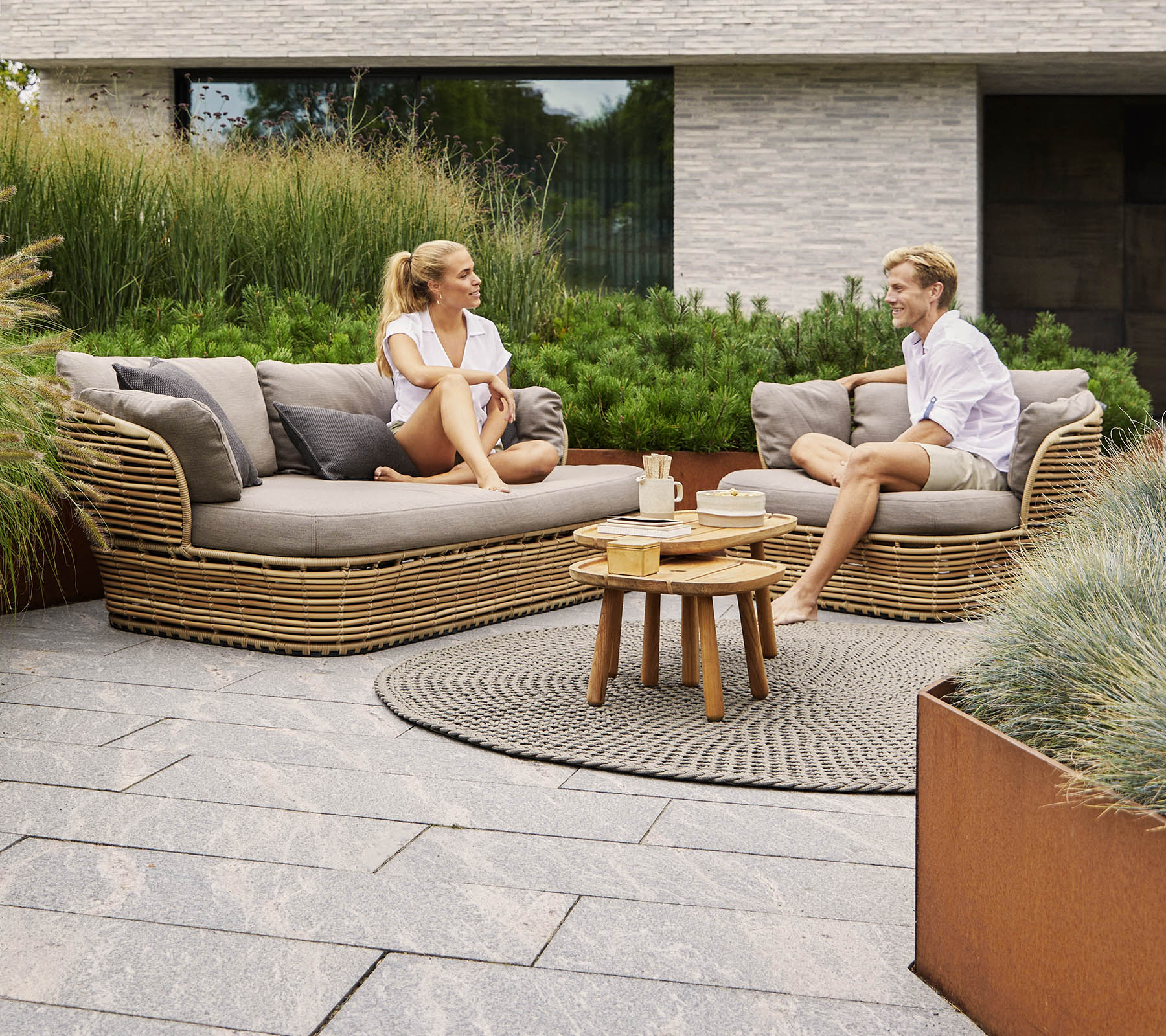 Two people converse while sitting on a stylish curved sofa, surrounded by greenery and modern decor, with a small wooden table between them.