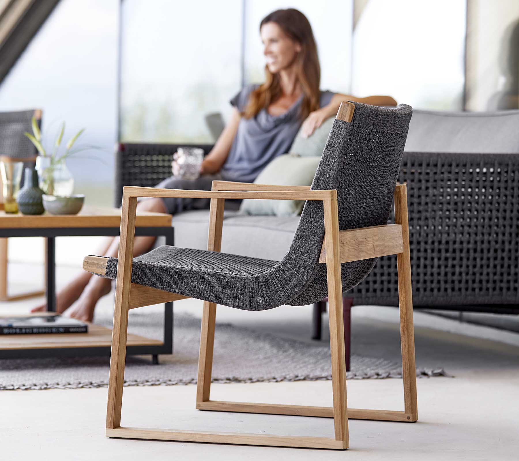 A modern chair with a sleek wooden frame and woven fabric, positioned next to a woman relaxing and enjoying a drink.