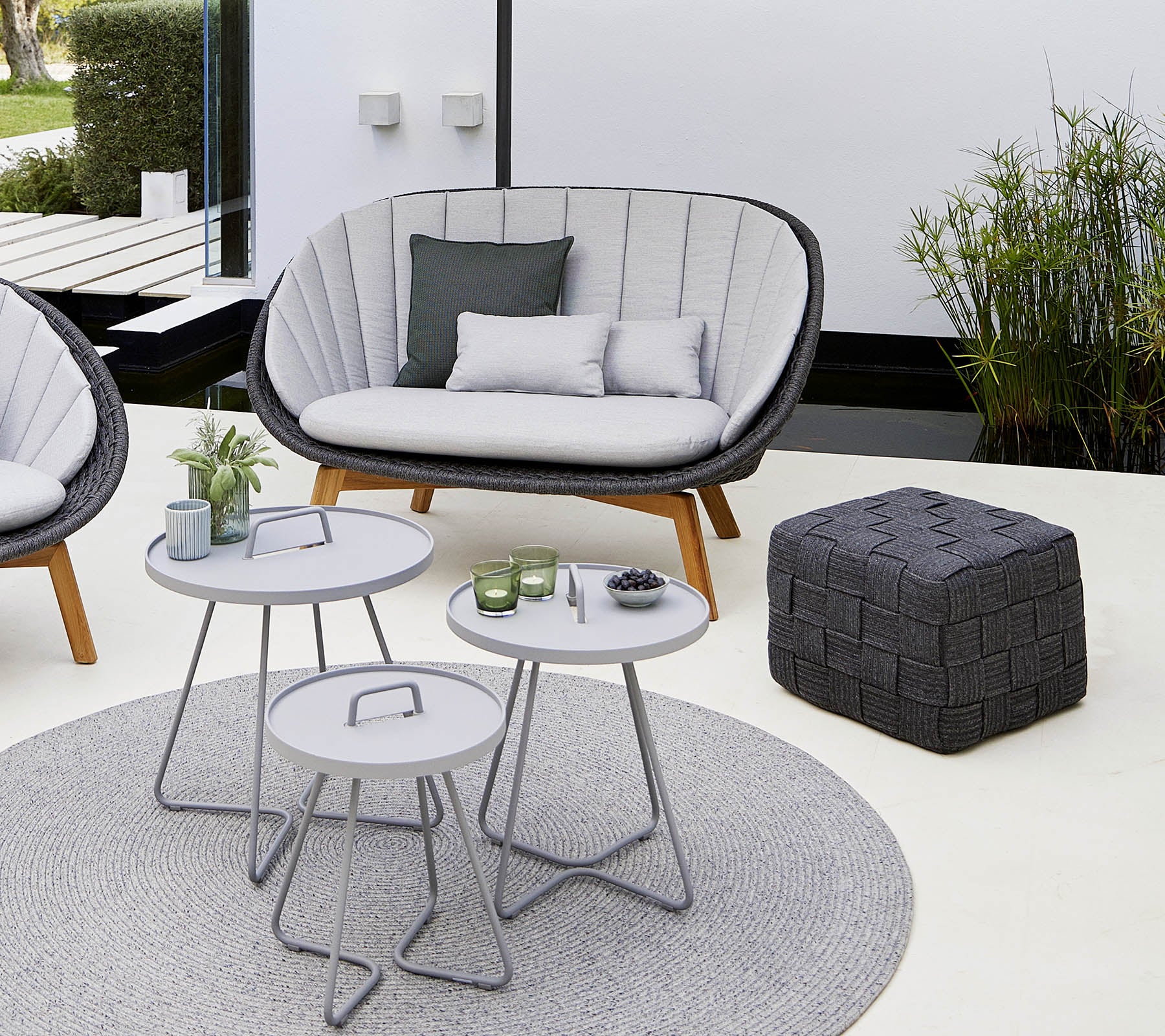 Cozy seating area featuring a stylish couch, a couple of round tables, a decorative pouf, and a soft rug, surrounded by greenery.
