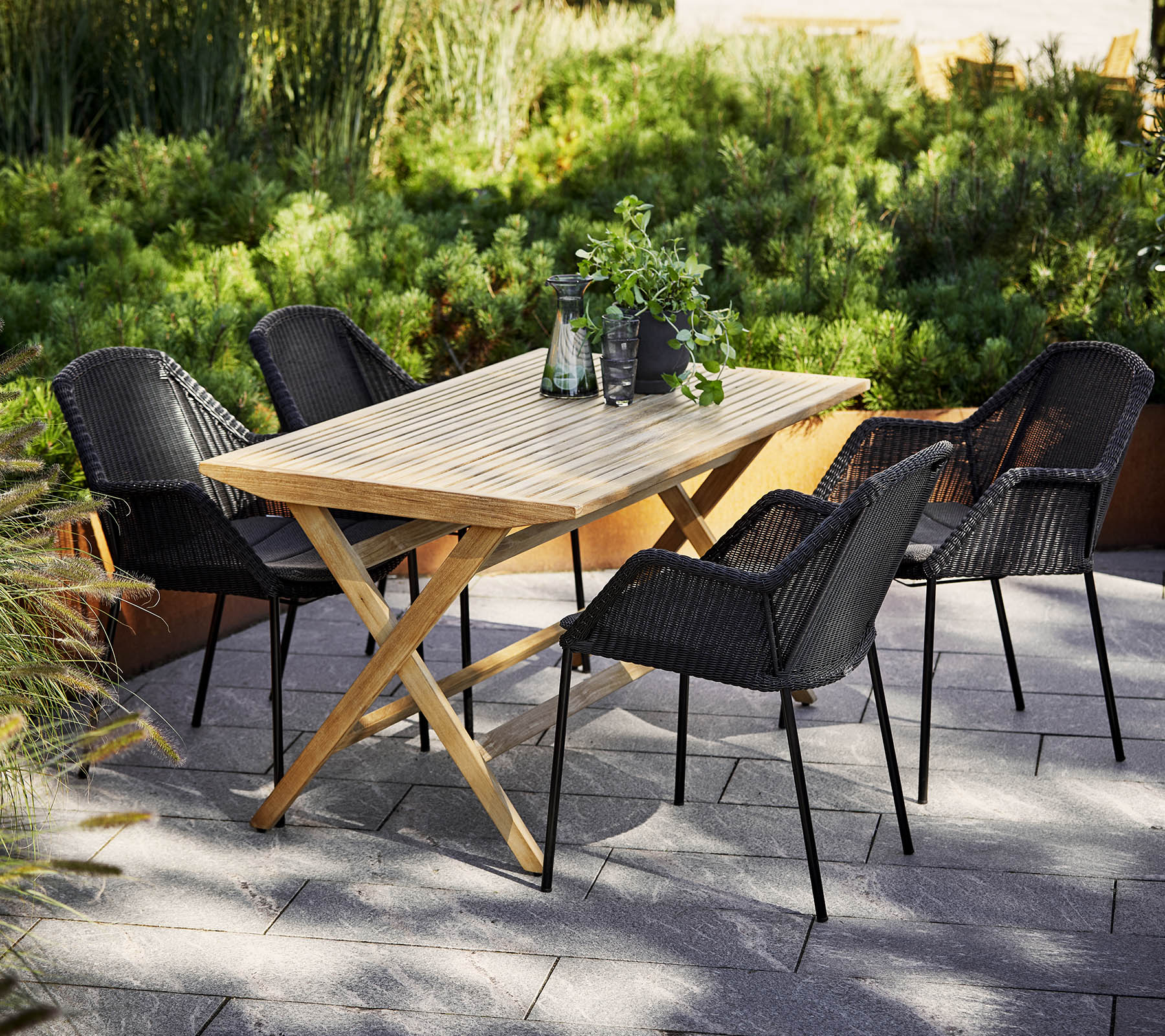 A wooden dining table surrounded by four black woven chairs, with greenery in the background and decorative items on the table.