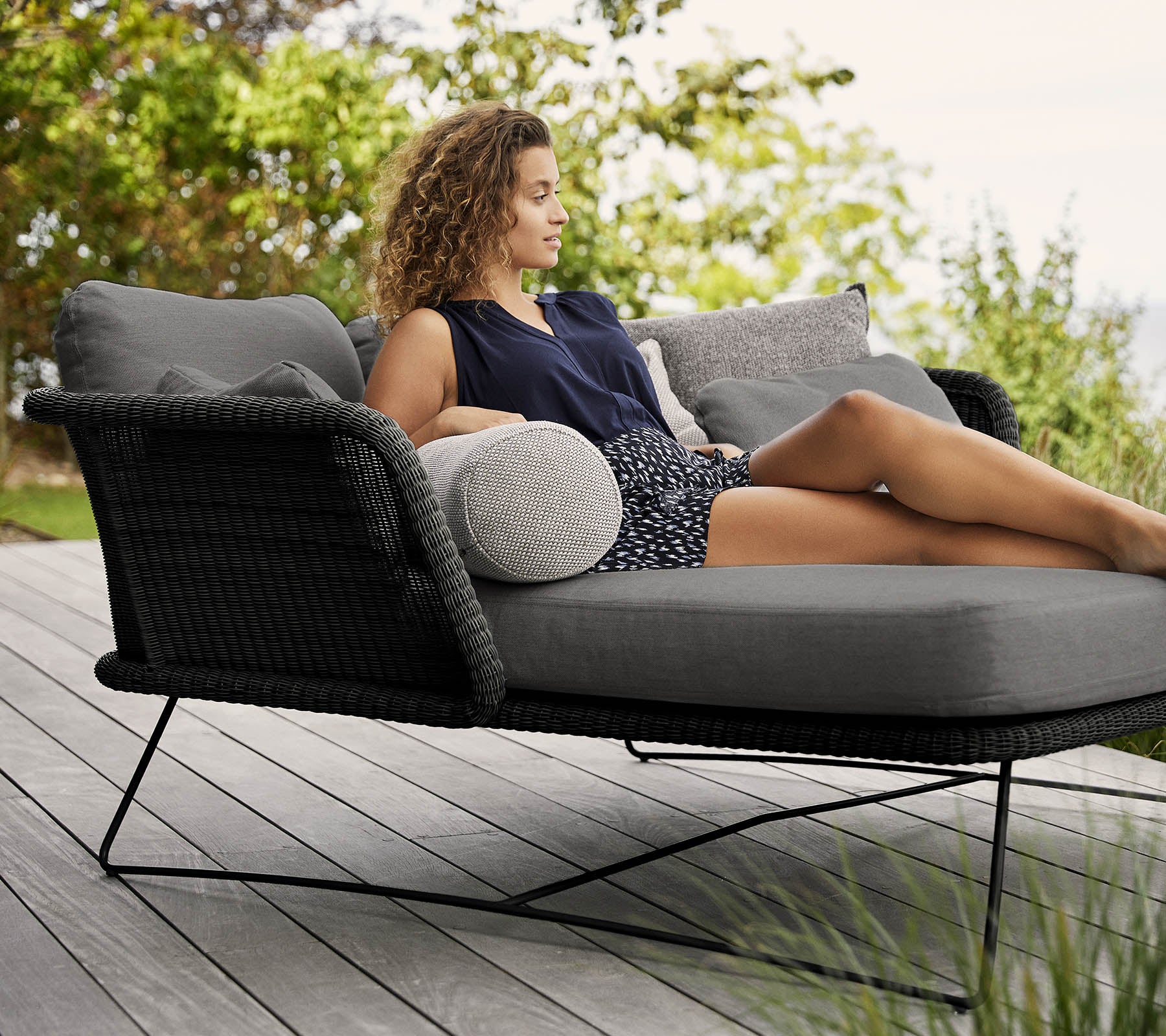 A woman relaxes on a stylish, cushioned lounge chair adorned with pillows, surrounded by greenery.