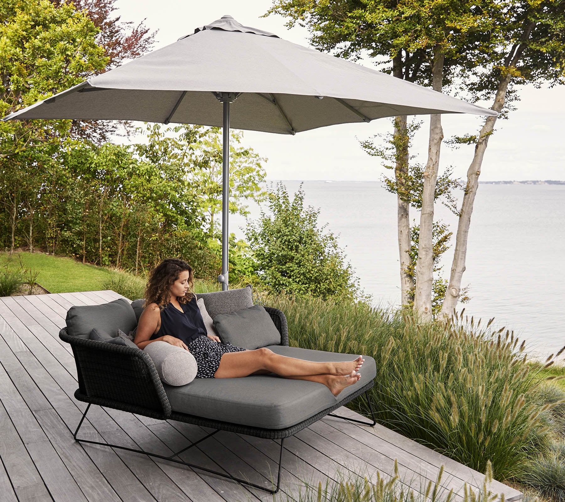 A woman relaxes on a modern lounge chair with a large umbrella, surrounded by greenery and a scenic waterfront view.