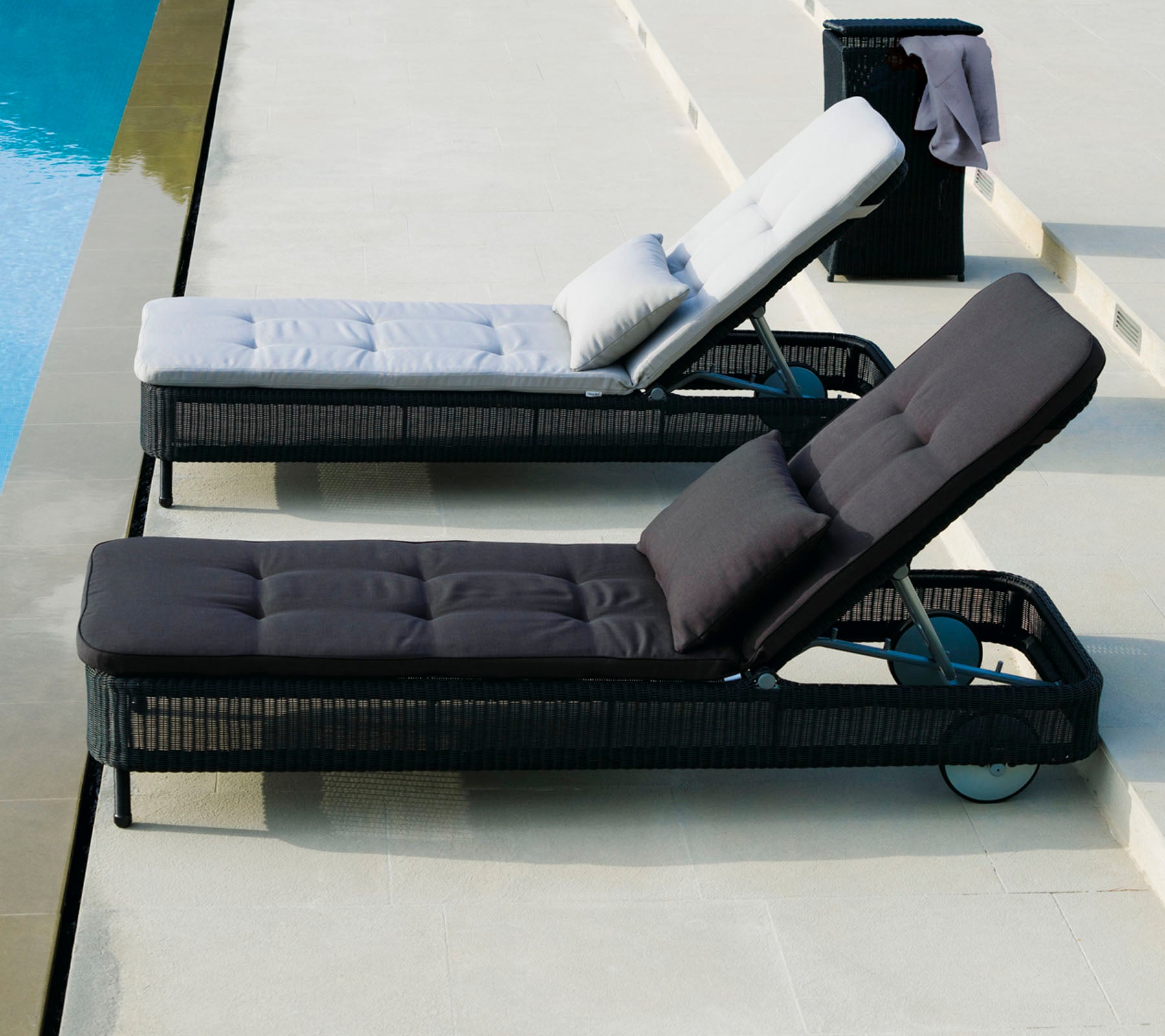 Two lounge chairs in contrasting colors, one in light gray with a pillow, and one in dark gray, positioned beside a pool.
