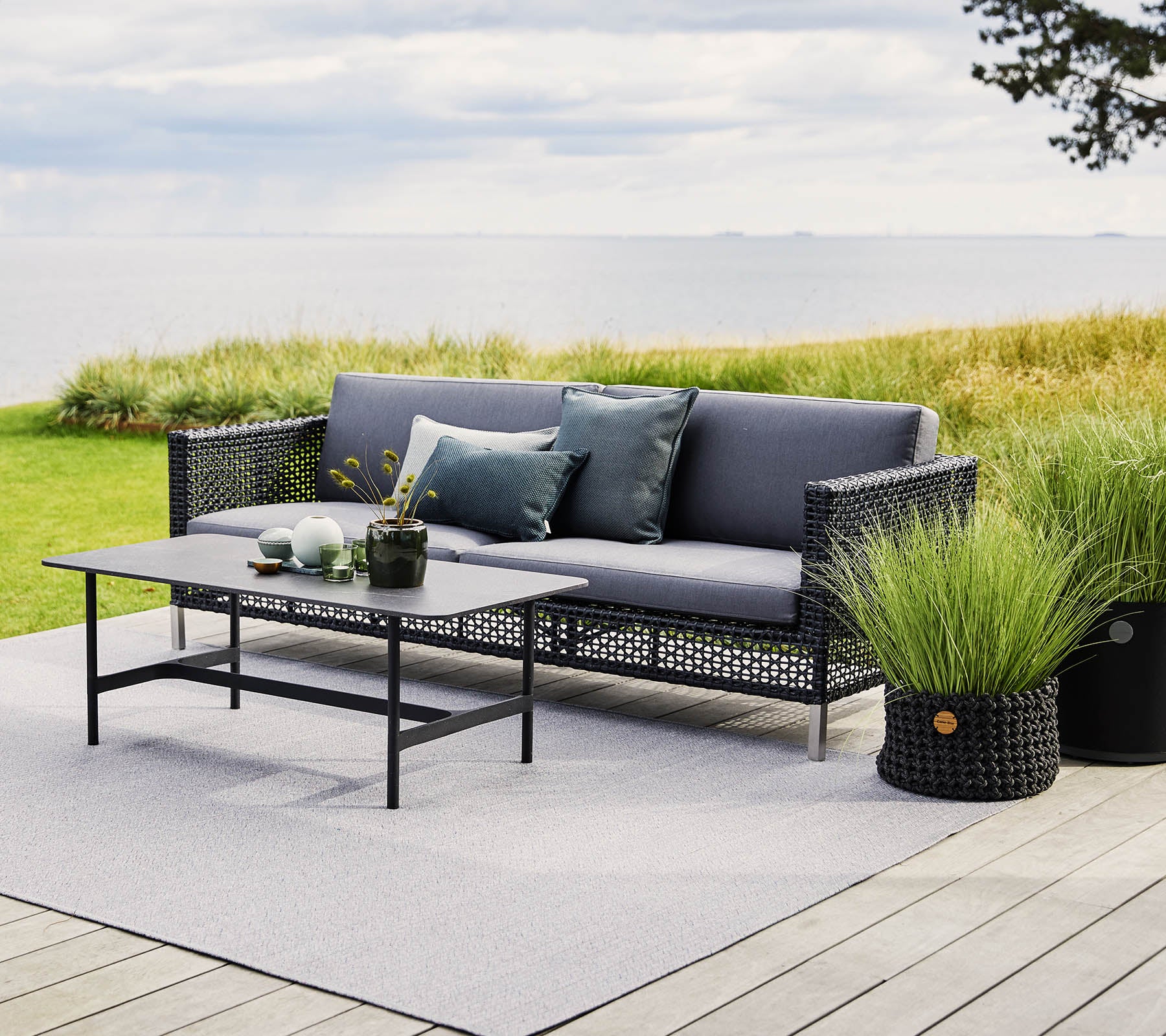 Stylish outdoor seating with gray cushions, a glass-top table, and potted plants, set against a scenic landscape.