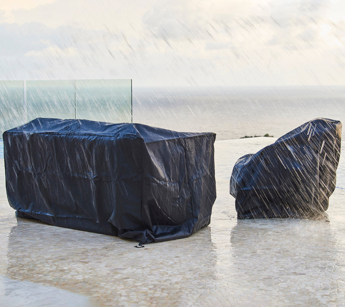 Outdoor furniture covered with dark protective covers, shielded from rain, with a backdrop of a cloudy sky and ocean.