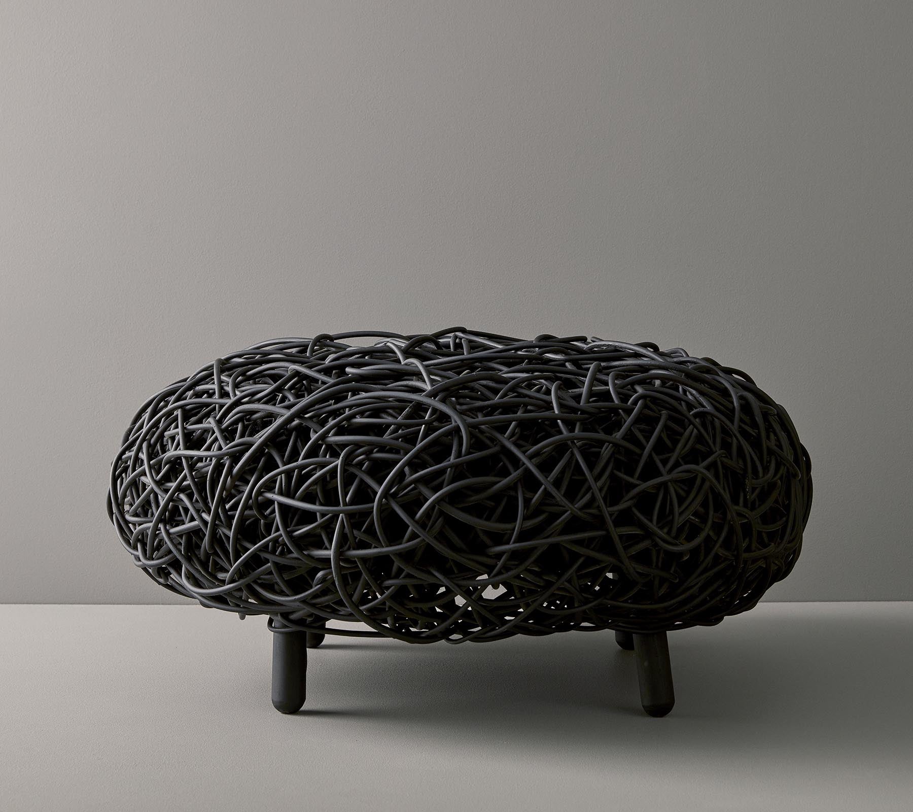 Black woven ottoman with an organic, rounded shape and intricate design, resting on three slender legs against a neutral background.