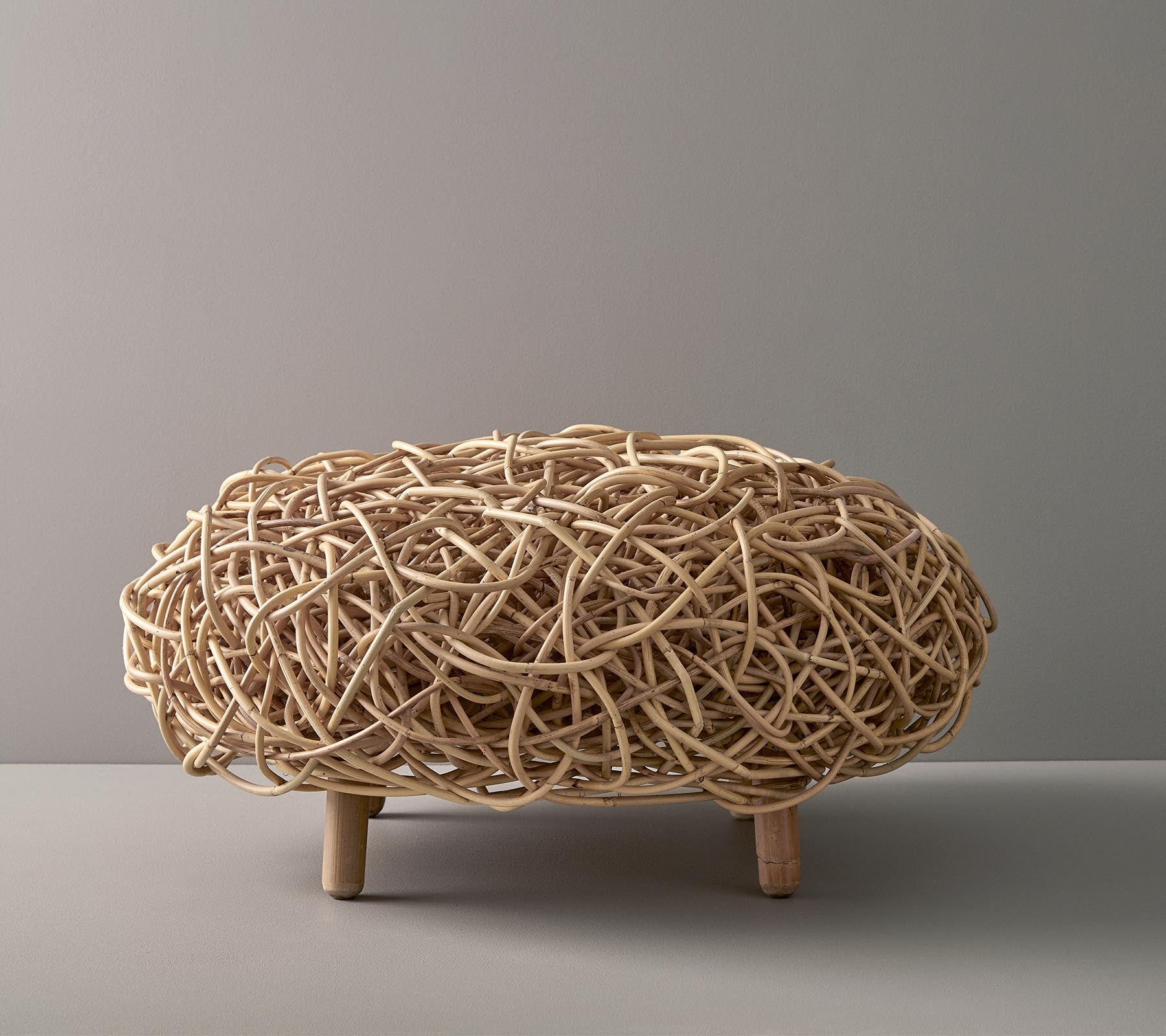 Natural woven decorative piece with an oval shape, featuring intricate interlaced strands and supported by short wooden legs.