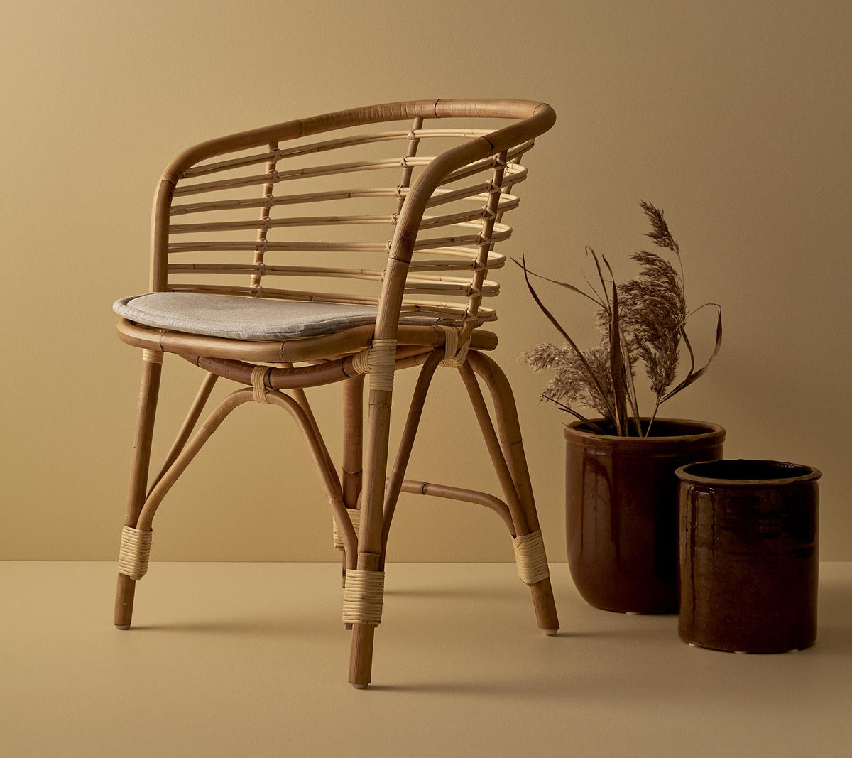 A stylish rattan chair with a cushioned seat, accompanied by two decorative planters featuring dried plants.