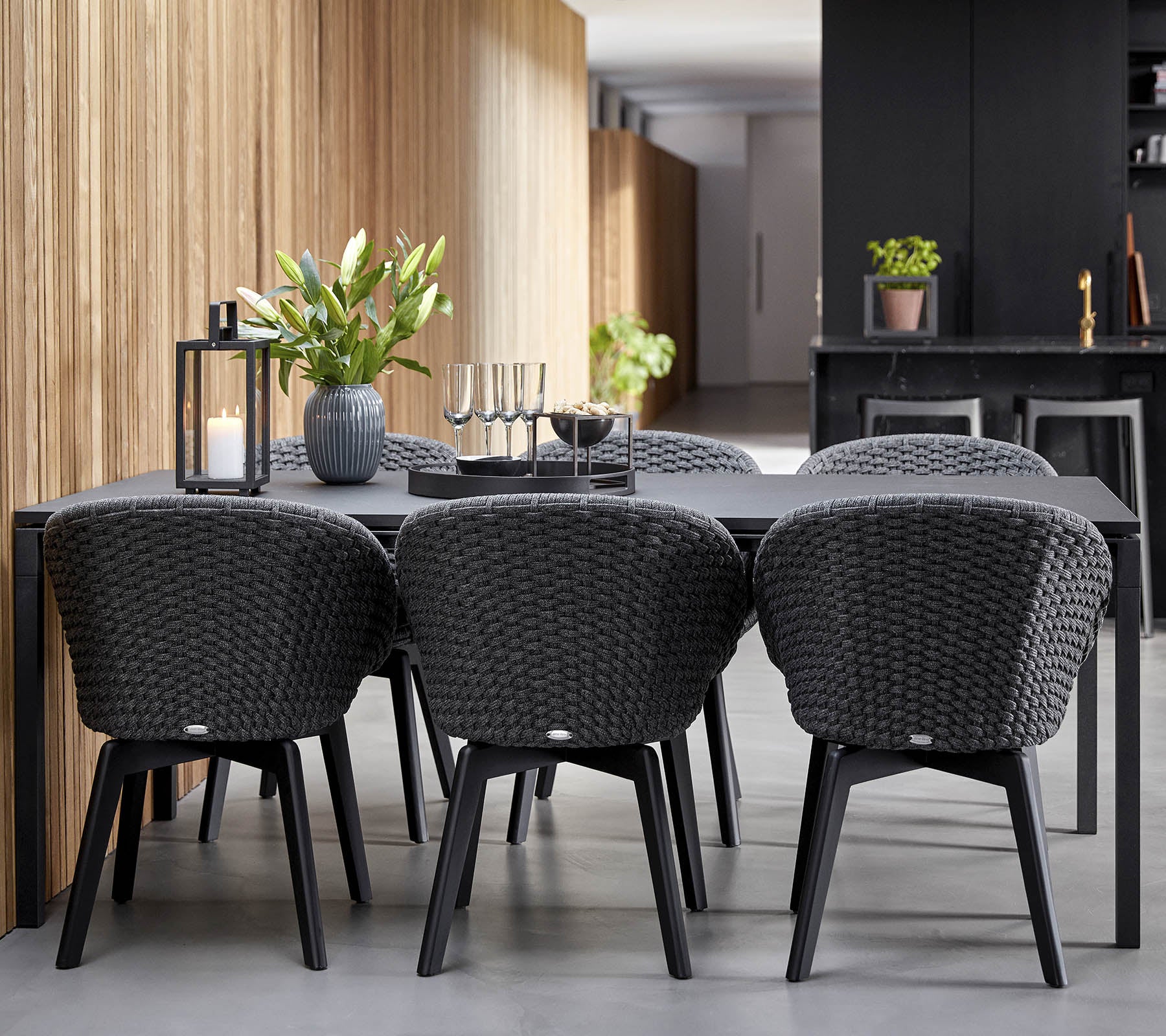 A modern dining setup featuring a sleek black table surrounded by stylish woven chairs, complemented by decorative plants and candles.
