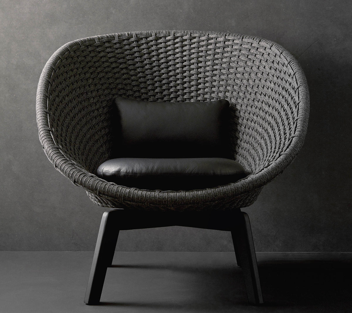 A stylish round chair with a textured woven design and a black cushion, resting on sleek wooden legs against a dark background.