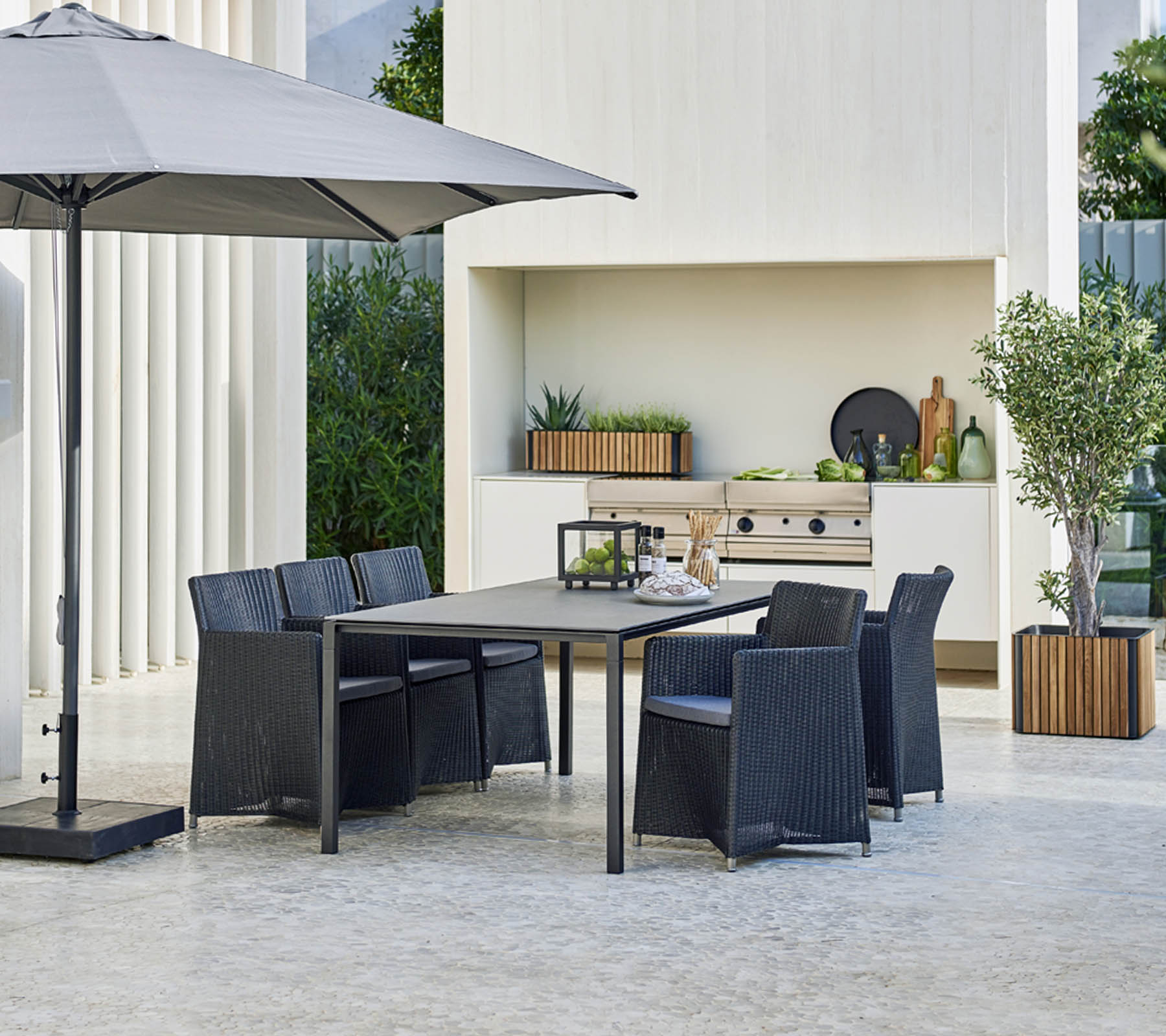 Modern outdoor dining set with a large table, six chairs, a stylish umbrella, and a backdrop featuring greenery and decorative elements.