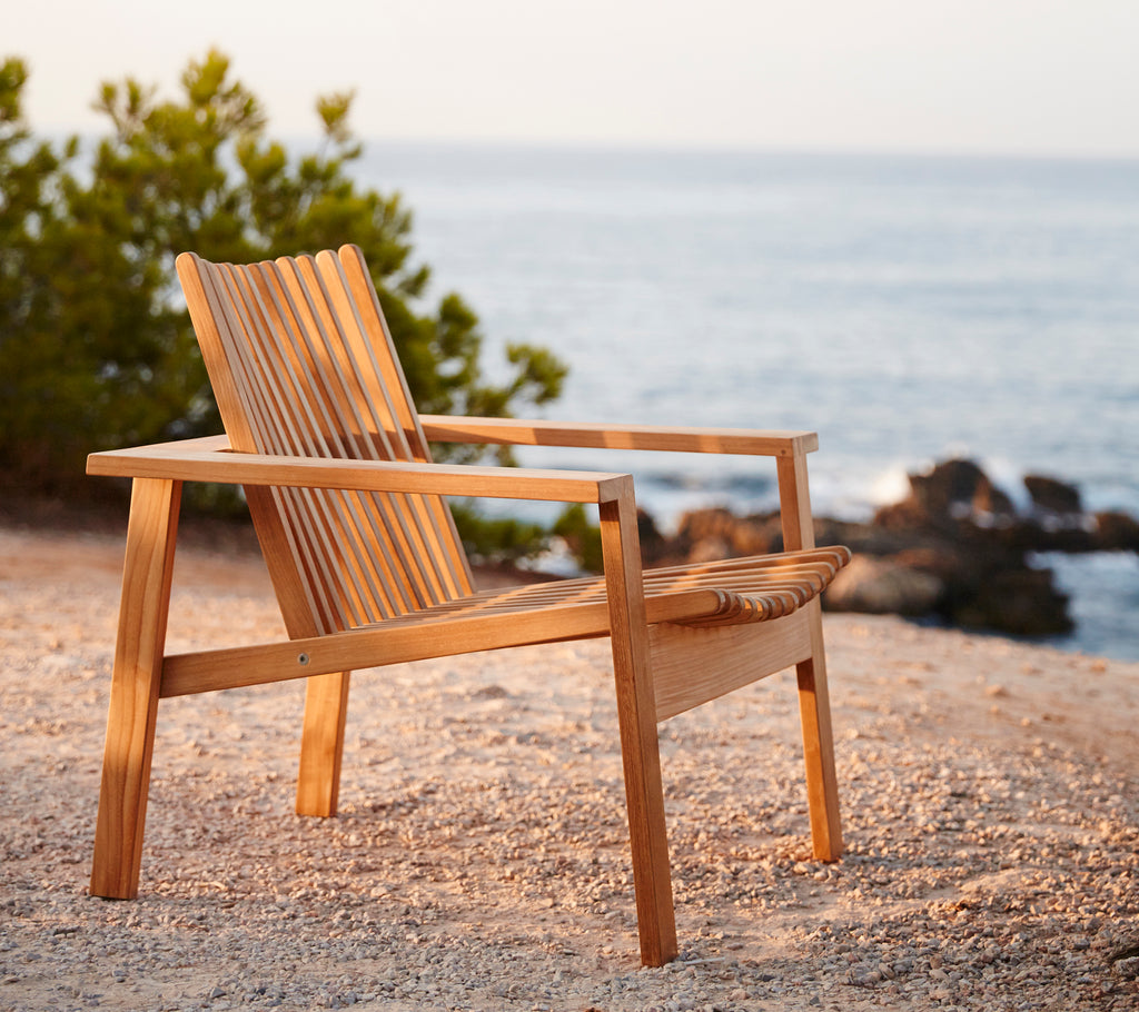 Side view of a wooden lounge chair with slatted backrest, positioned near a serene water scene and surrounded by greenery.