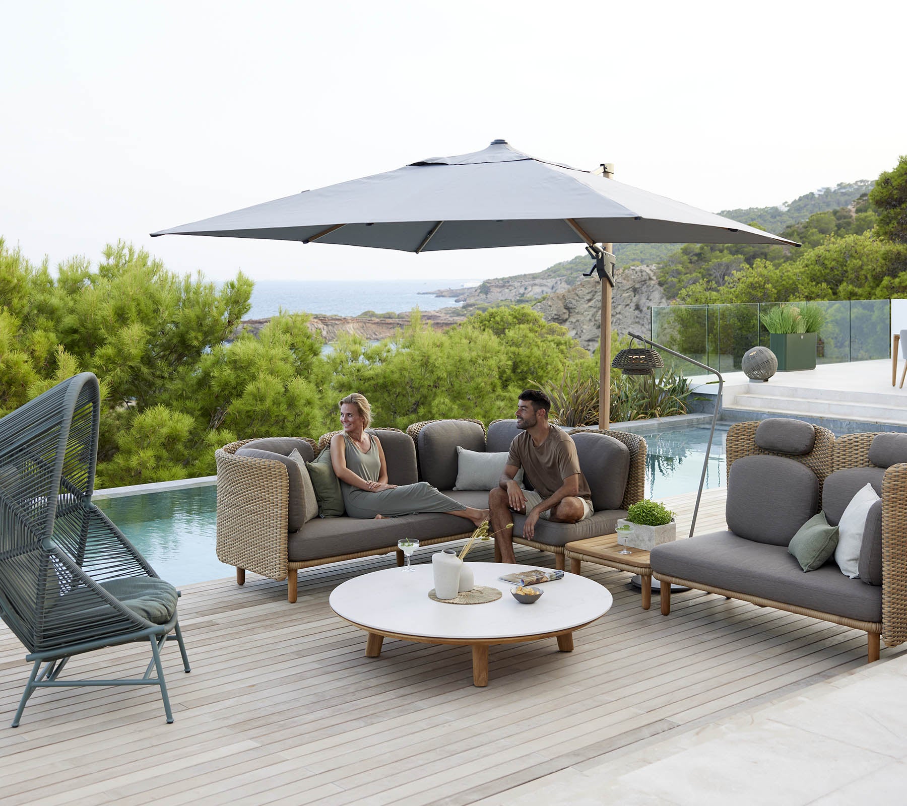A stylish lounge area featuring comfortable seating, a large umbrella, and a round table, surrounded by greenery and a serene view.