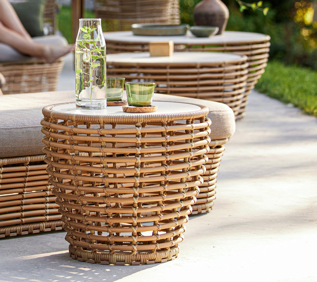 Small basket coffee table featuring a woven design with a glass top, accompanied by a water bottle and green glassware.