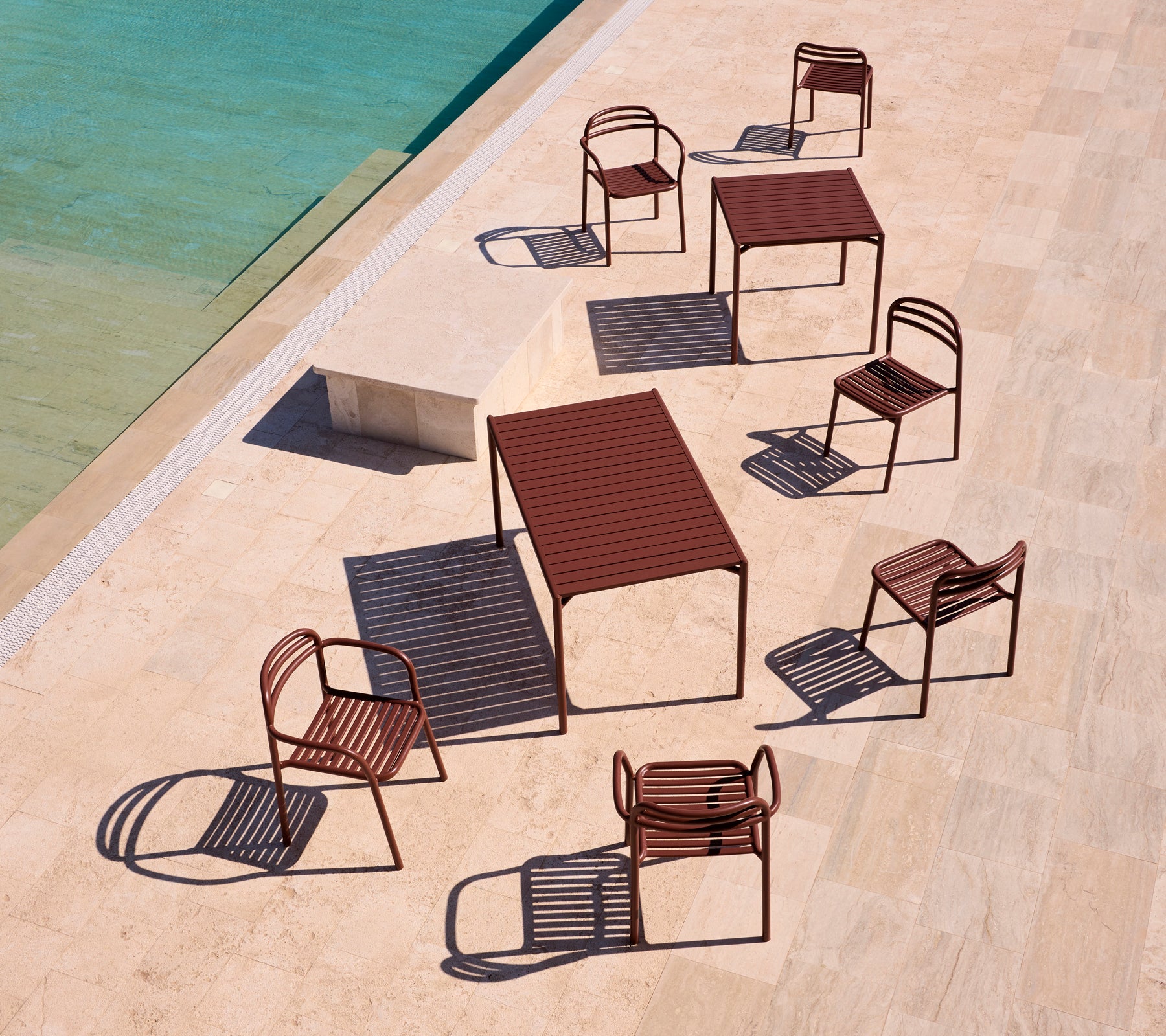 Bliss series outdoor furniture in desert red, arranged around a pool on a sunlit terrace, casting playful shadows.
