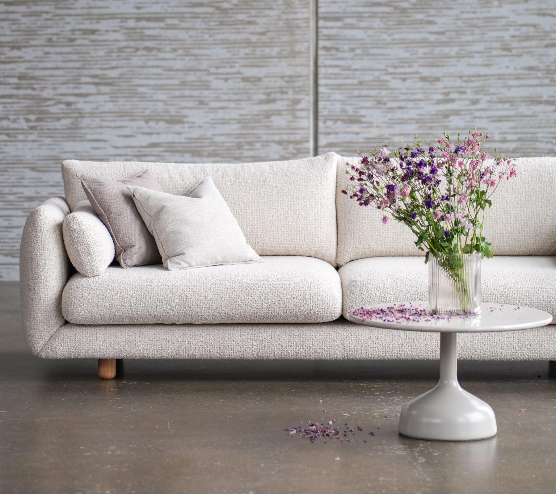 Light-colored sofa with teak legs, adorned with cushions, complemented by a round table and a vase of fresh flowers.