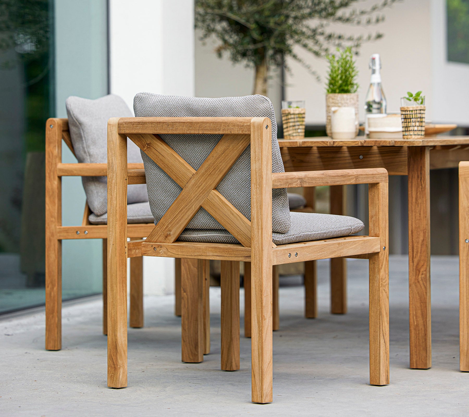 Wooden chairs with gray cushions and a matching table, accompanied by decorative plants and a bottle, ideal for outdoor dining.