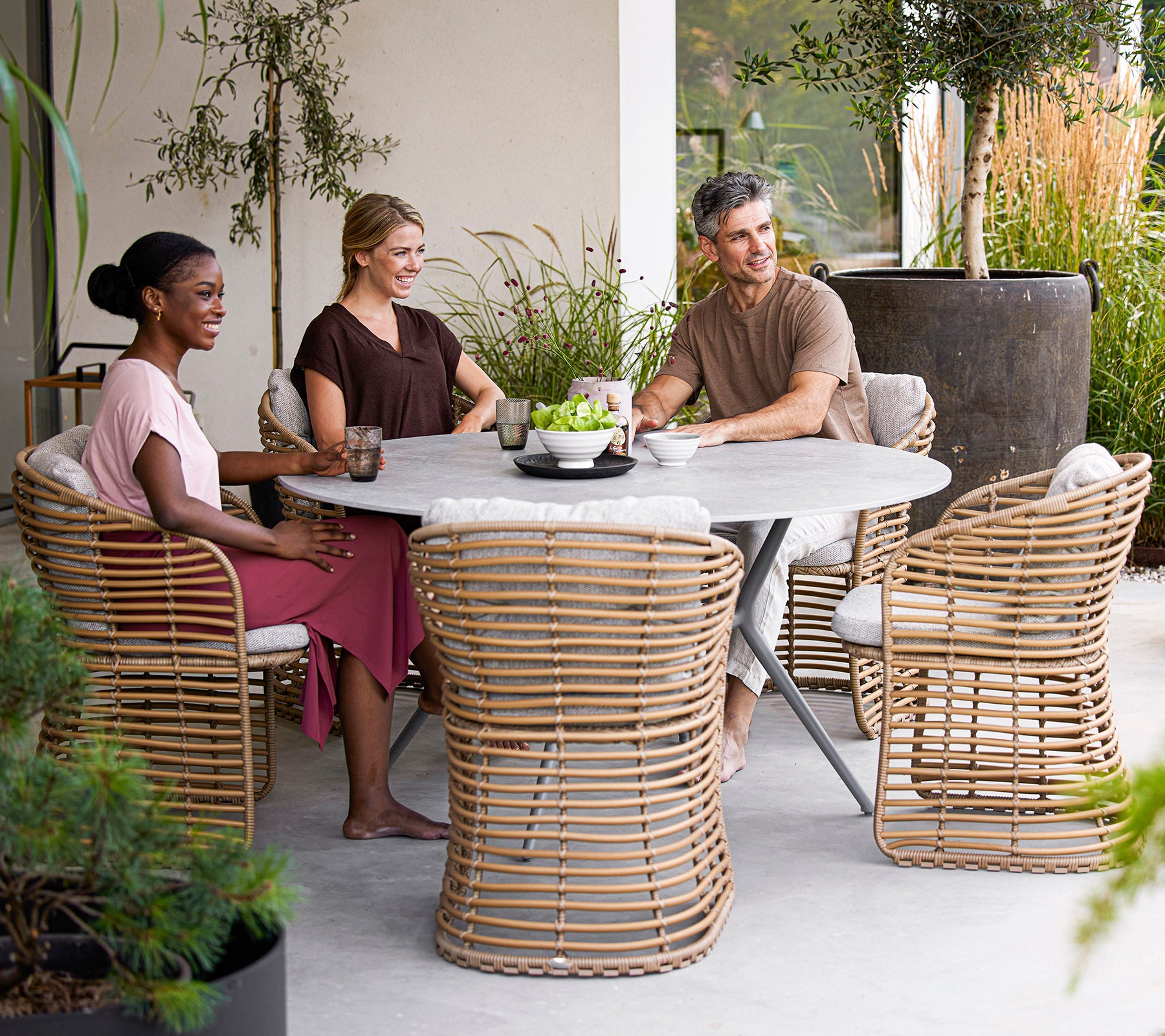 Four people enjoy drinks around a modern round table, with stylish rattan chairs and greenery in a serene setting.