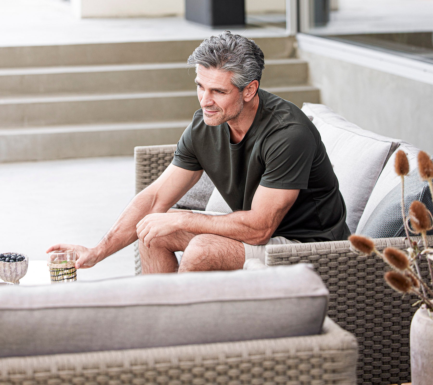 A man sits casually on a sofa, holding a drink and smiling, surrounded by soft cushions and decorative elements.