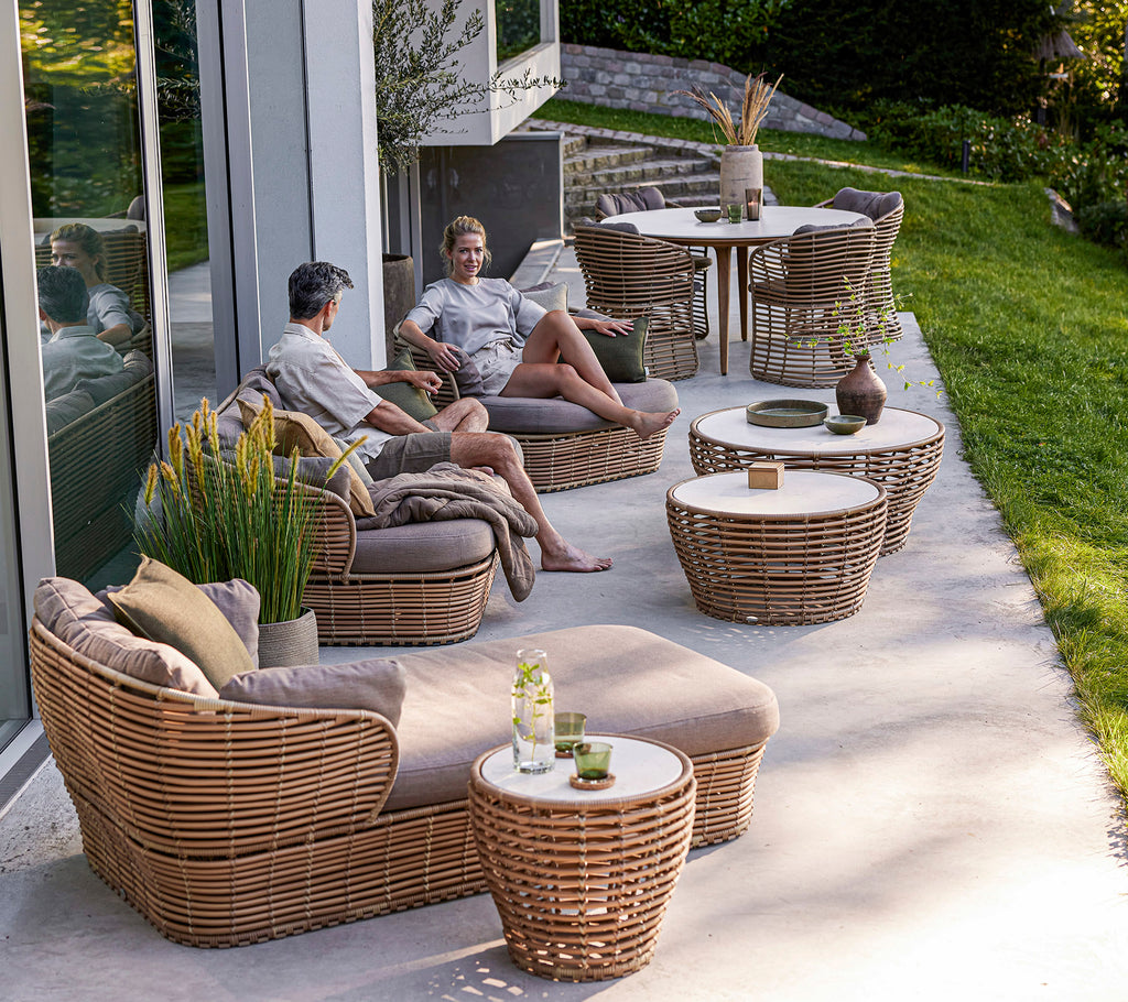 Two individuals relax on woven furniture surrounded by tables and greenery, enjoying a casual outdoor setting.