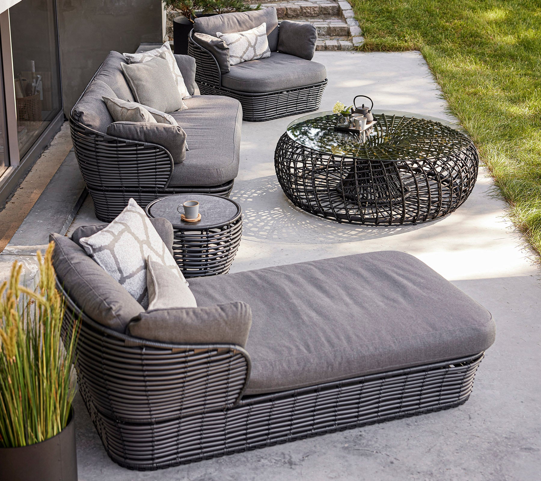 Stylish outdoor seating arrangement featuring woven furniture, cushions, a glass coffee table, and decorative plants. Ideal for relaxation.