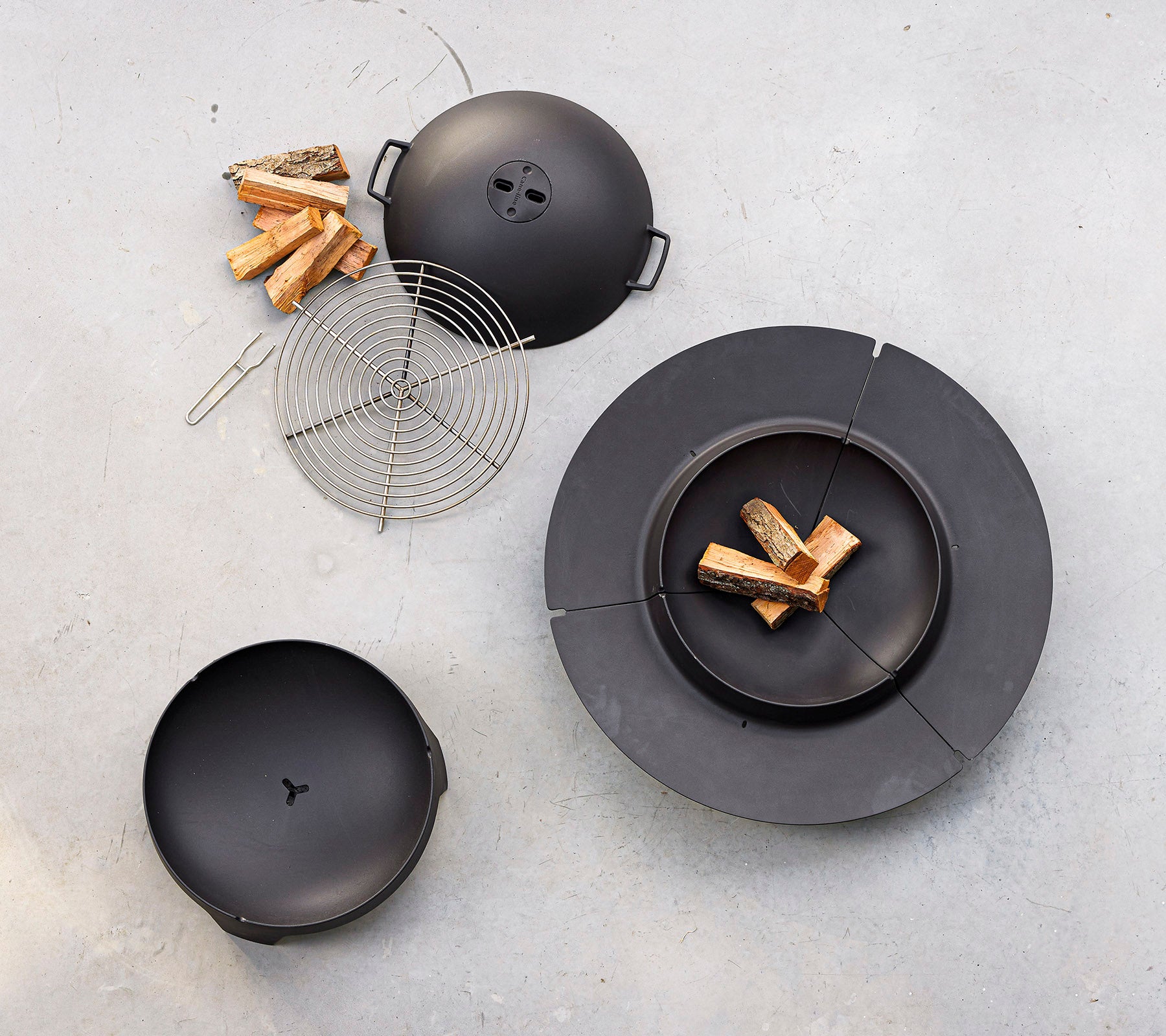 A black cooking set featuring a circular grill with a lid, a grilling rack, and firewood pieces arranged nearby.