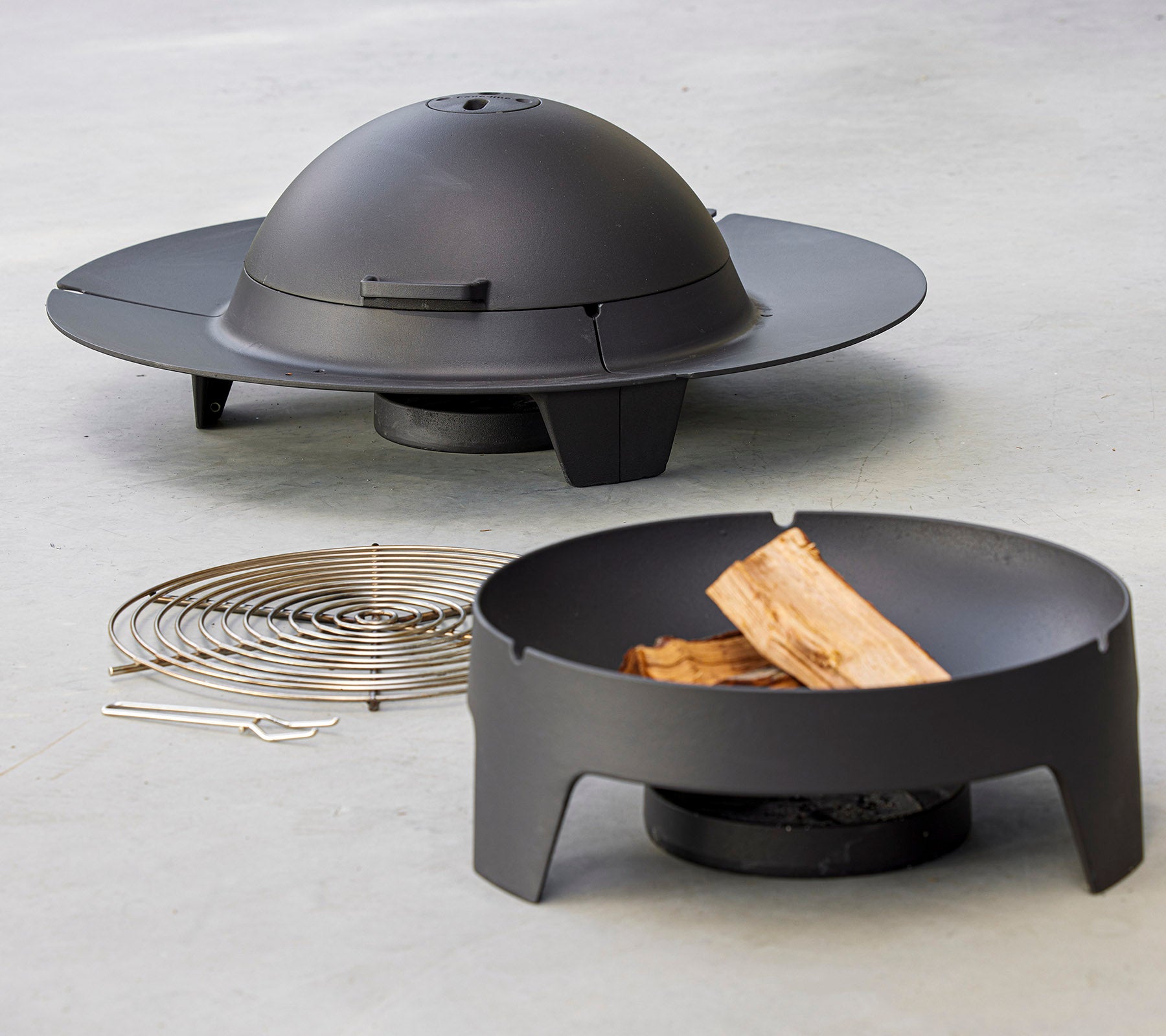 A modern firepit set featuring a sleek, circular design with a dome lid, accompanied by a grill rack and wood pieces.
