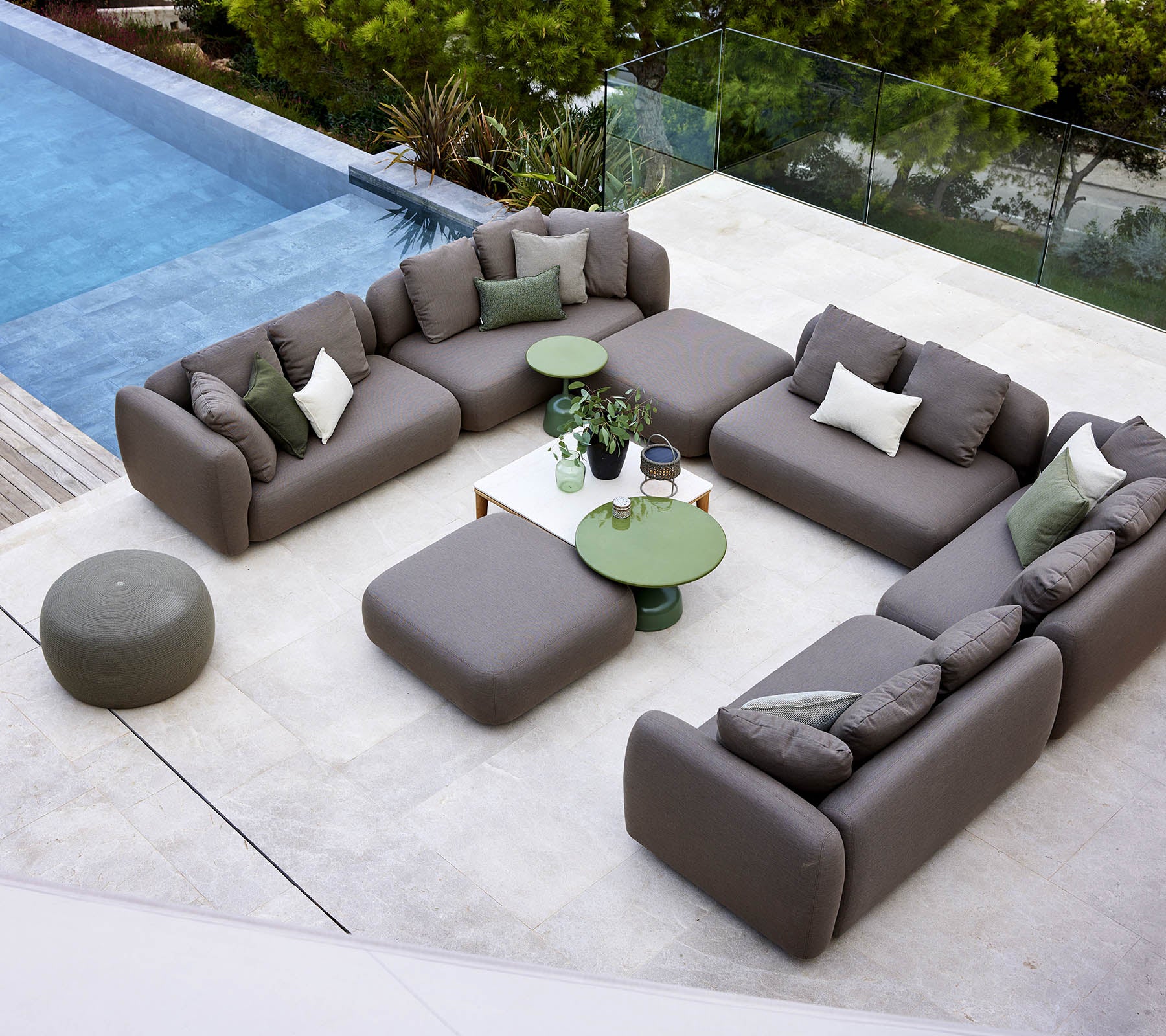A modern lounge setup featuring a sectional sofa, round ottoman, and green accent tables, surrounded by greenery and a tranquil pool.