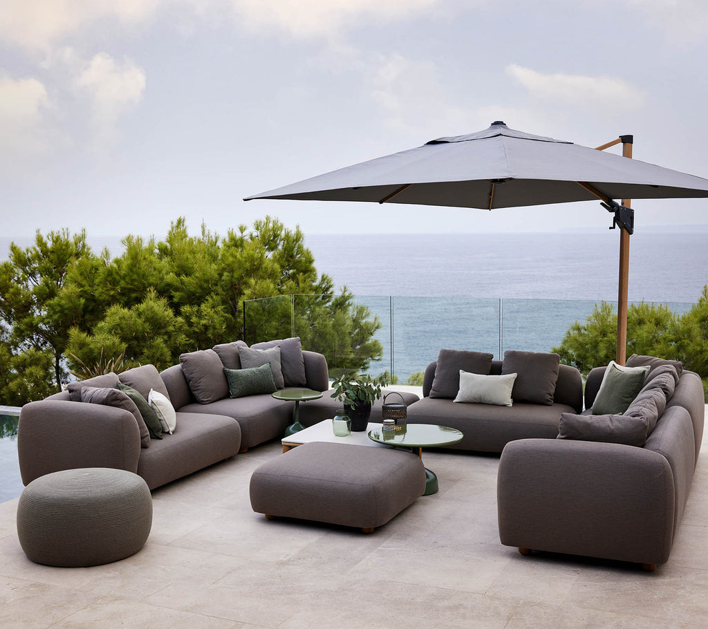Modern lounge seating set with plush cushions, accompanied by a round table and a large umbrella, surrounded by greenery and a scenic view.